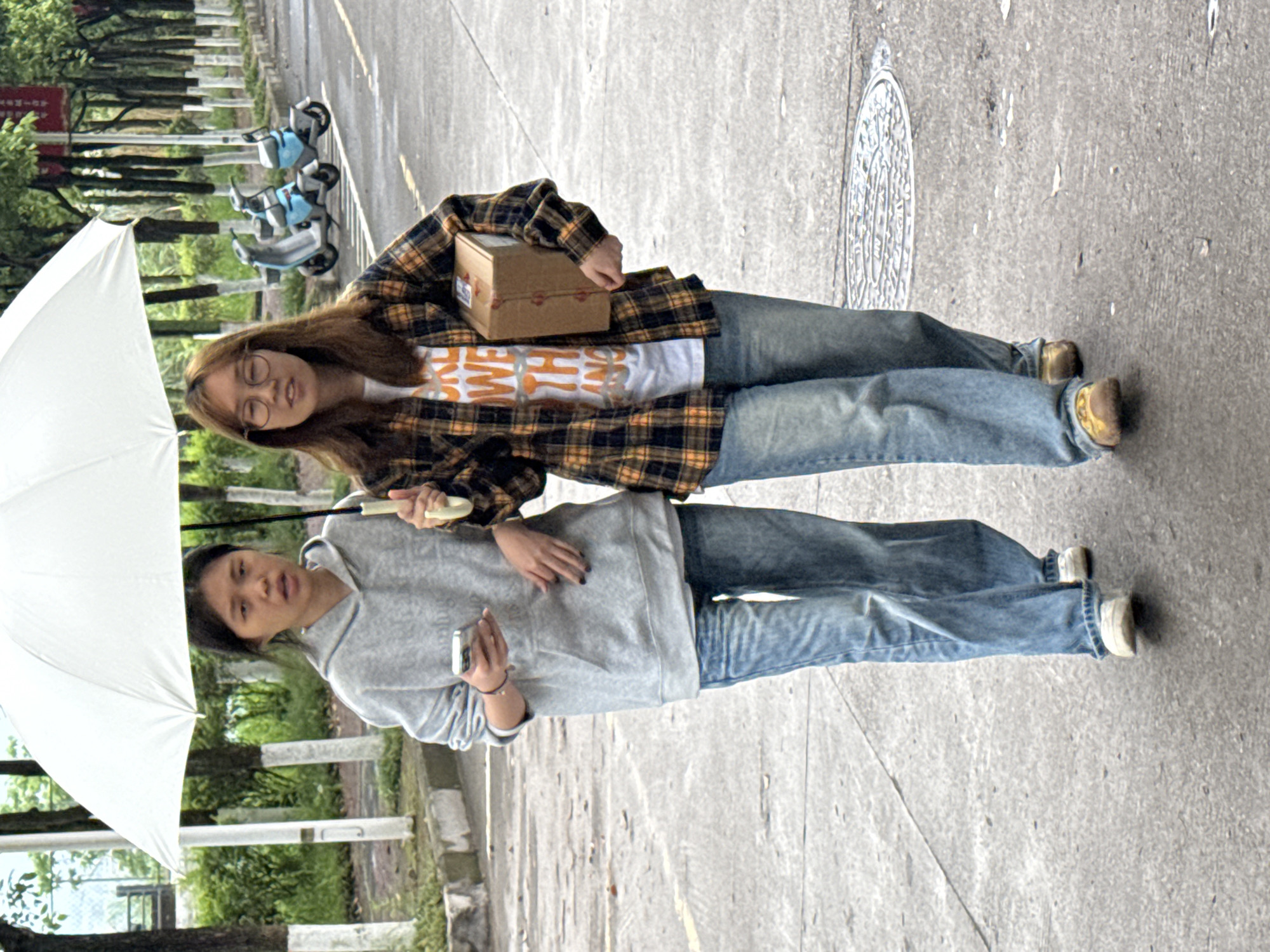 Two women stand shoulder-to-shoulder on a paved road under a white umbrella. One woman wears glasses, a plaid shirt over a printed t-shirt, and holds a small brown box. The other woman wears a gray hoodie and holds a mobile phone. The location is likely in Jiangjin, China, evidenced by the presence of parked scooters in the background and general urban landscape.