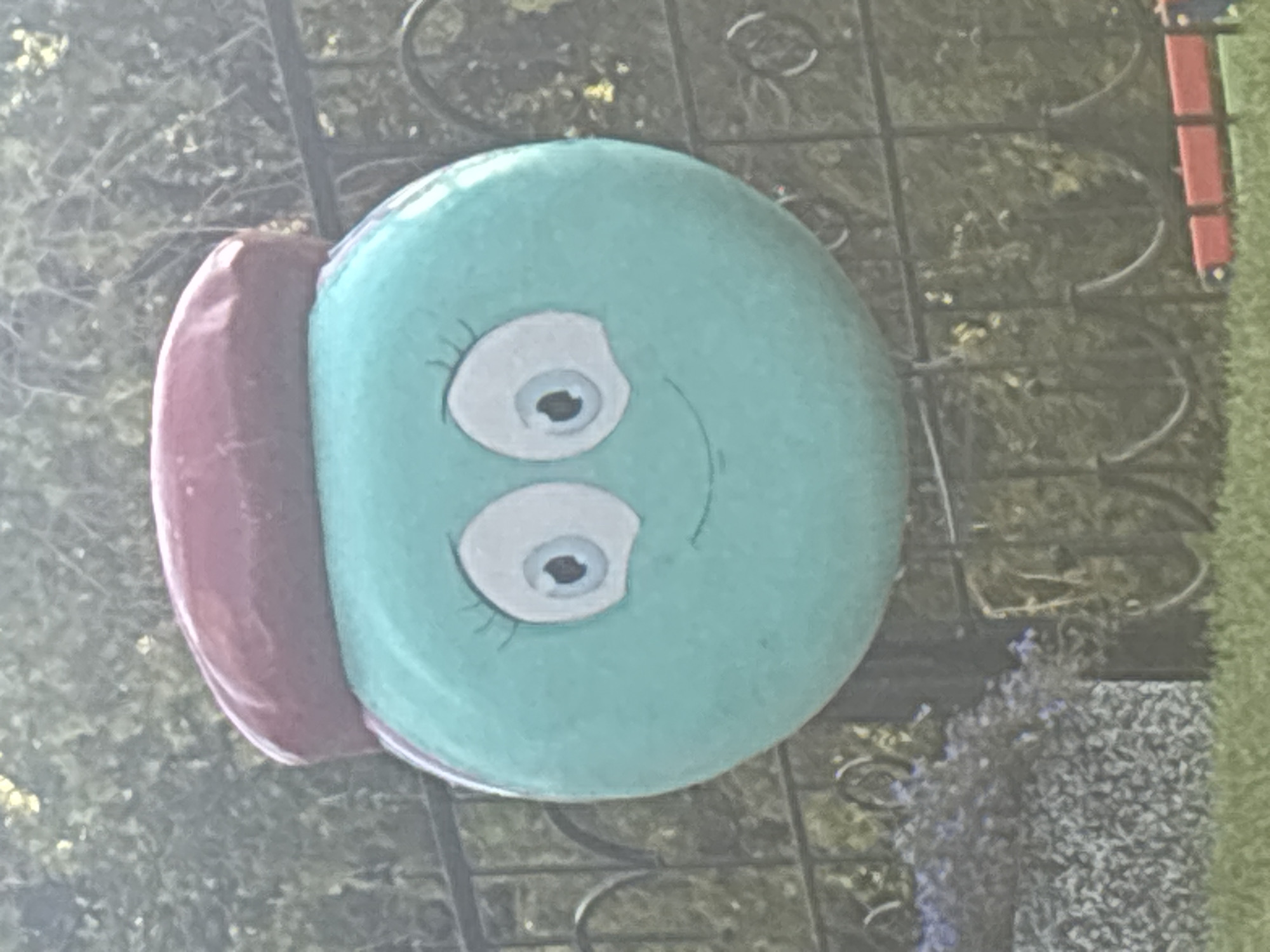 The image shows a close-up of a round, teal-colored garden ornament with a painted cartoon face. The face has large white eyes with black pupils, long eyelashes, and a smiling mouth. A rounded, red beret-like hat sits on top of the ornament. The ornament is positioned in front of a black wrought-iron fence, with blurred green foliage and trees in the background. In the lower right corner, a portion of a red and green object, possibly a toy or another garden decoration, is visible. The ground in the foreground is covered in green grass. The image does not contain any information that would allow for its specific location in Novosibirsk, Russia.
