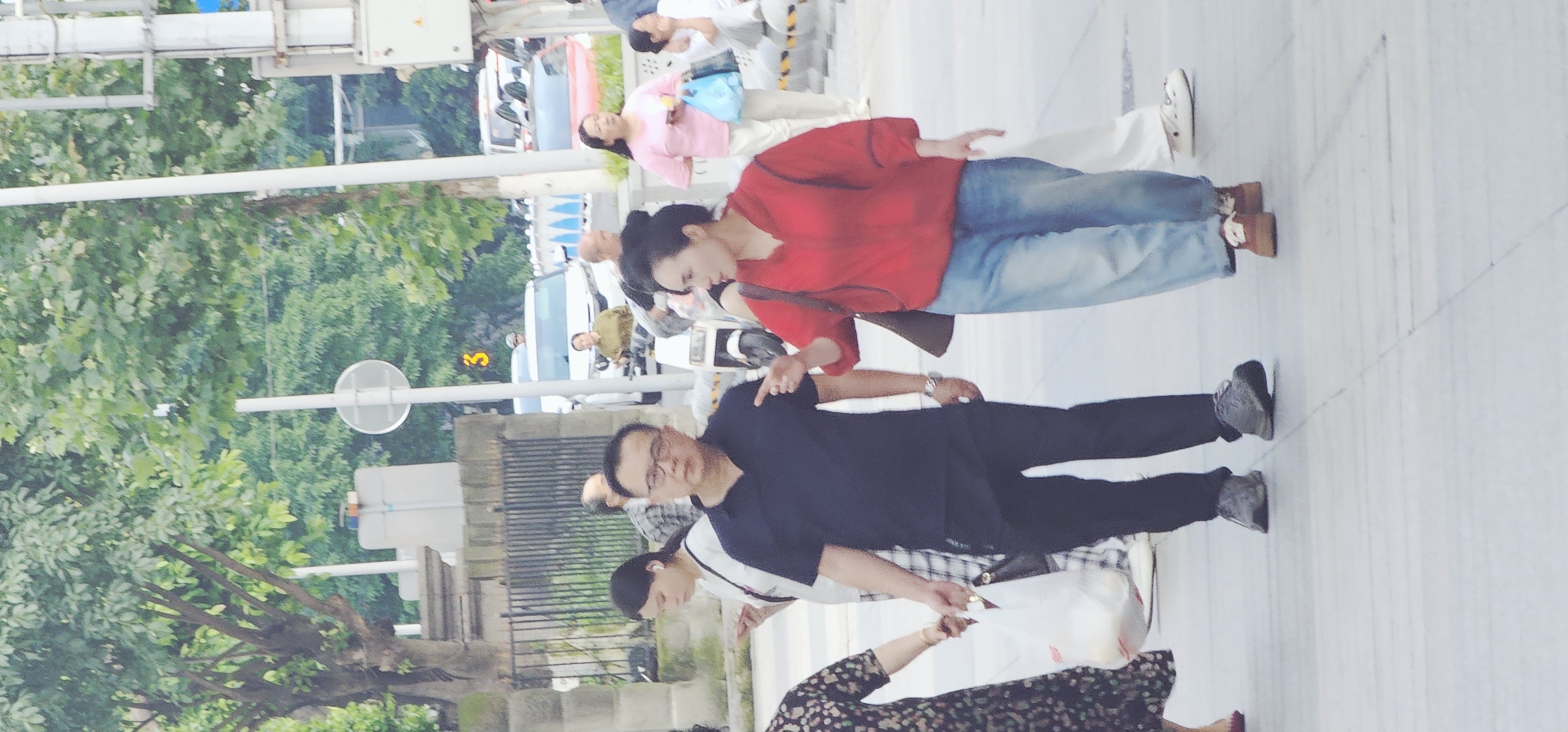 A medium shot taken in the Yuzhong District of China captures a bustling street scene. The focus is on several pedestrians walking on a paved walkway. A man in a dark blue shirt and black pants is walking towards the camera, while a woman in a red sweater and jeans is beside him. They appear to be on a busy thoroughfare with trees and buildings visible in the background. There are also other people walking in the vicinity, with cars parked along the street. The scene gives the impression of a typical day in a densely populated urban area.