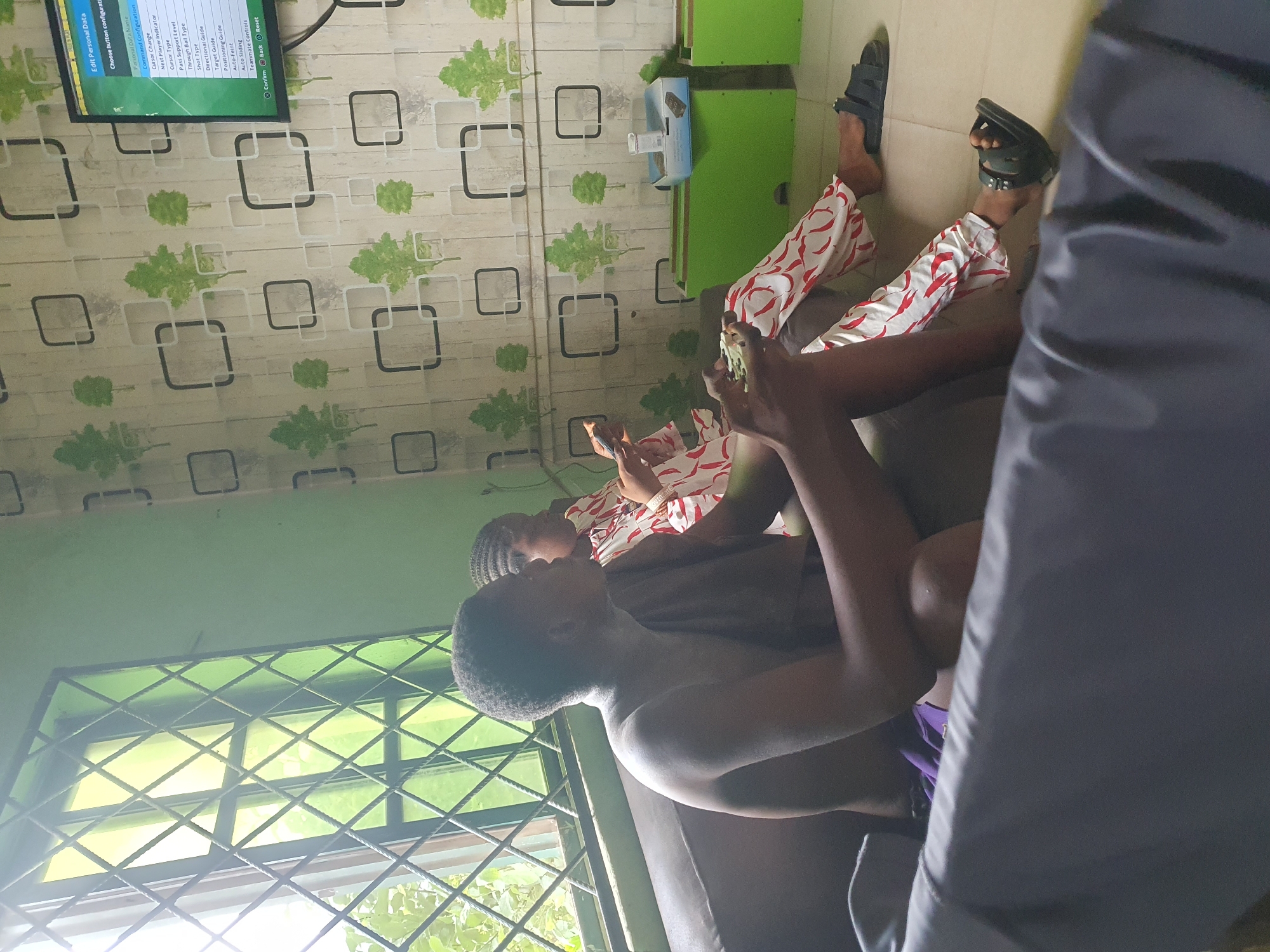 Here is a description of the image:
The image shows two young people in a room, presumably in a residential setting in Lokoja, Nigeria. One person, wearing patterned red and white clothing, is playing a video game on a flat screen television mounted on the wall. The TV displays game settings. The second person is shirtless. Both people are sitting on a couch. Beside the couch there's what appears to be a green cabinet with a fan on it. The room has a window with a diamond patterned security bars. The wall has a patterned wallpaper with green leaf designs. The image is shot from a low angle, and a person wearing a dark blue garment appears on the right side.