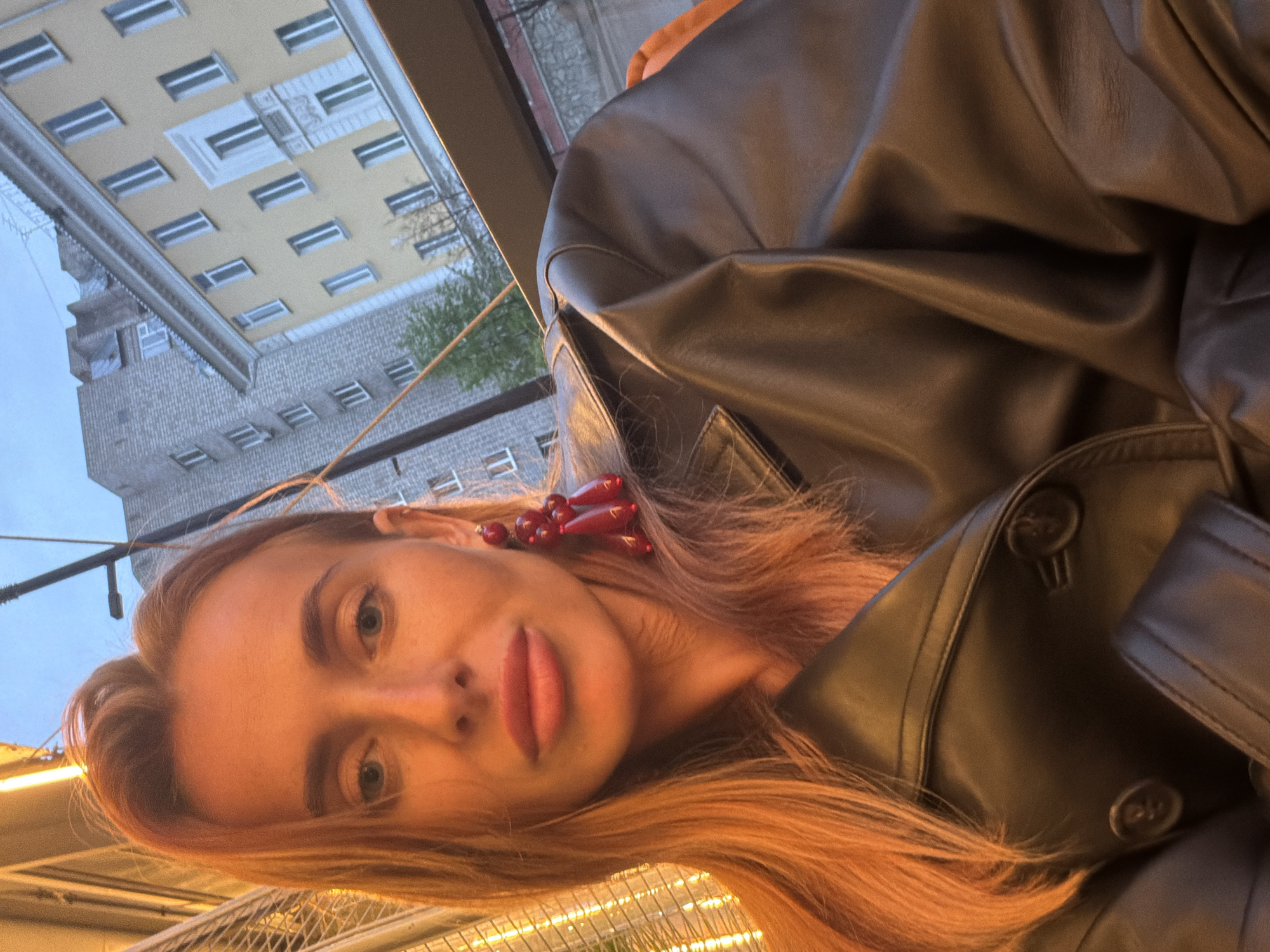 A woman with long, auburn hair is wearing a black leather jacket and red earrings. She is sitting outside, possibly at a restaurant. The background shows a building with many windows, possibly an apartment building. The location is likely in Krasnoyarsk, Russia, judging by the style of the buildings and the person's appearance.