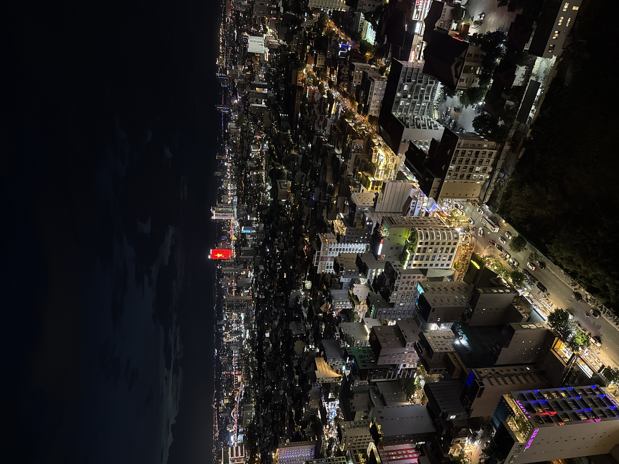 This high-angle night photograph captures a sprawling coastal city, offering a panoramic view of the illuminated urban landscape stretching horizontally across the frame. To the left, the city borders a vast, dark body of water, likely the sea, with a string of lights marking the coastline. The densely packed cityscape is a mosaic of countless lights emanating from buildings, streets, and vehicles, signaling a vibrant and active metropolis.
A prominent red banner or flag adorned with a yellow star, unmistakably the national flag of Vietnam, stands out on a structure located along the coastline, confirming the location as Vietnam. Given the context, this is likely Da Nang, a major coastal city.
The foreground, towards the right side of the image, reveals more detailed architecture, including multi-story buildings and visible streets with moving traffic. On one building, the text "PALAZI HOTEL" is clearly visible, identifying a commercial establishment. Other buildings exhibit distinct lighting, such as blue and red accents. The sky is dark, indicative of nighttime, with faint cloud formations discernible over the water. The overall scene conveys the lively atmosphere of a bustling city continuing its activities under the night sky.