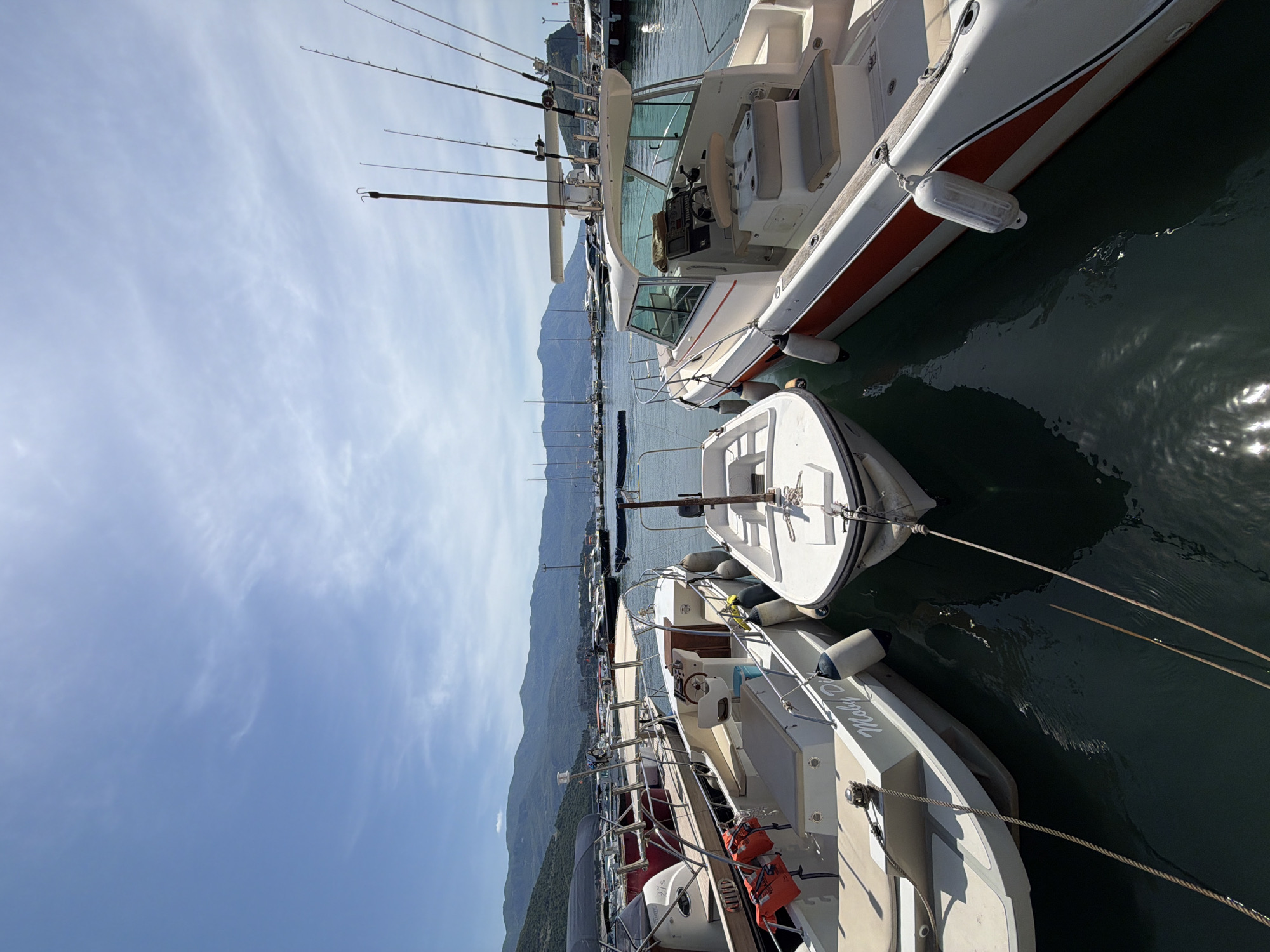 The image shows a marina with several boats docked, likely in Budva, Montenegro, given the mountainous backdrop and coastal setting. The foreground features multiple boats of varying sizes and designs, some equipped with fishing rods. The boats are moored close together, with ropes visible connecting them to the docks. The water is calm, reflecting the sky, and the sky itself is partly cloudy. In the distance, there are more boats and the outline of hills or mountains, a common feature of the Montenegrin coastline. The overall impression is of a peaceful marina scene on a sunny day.