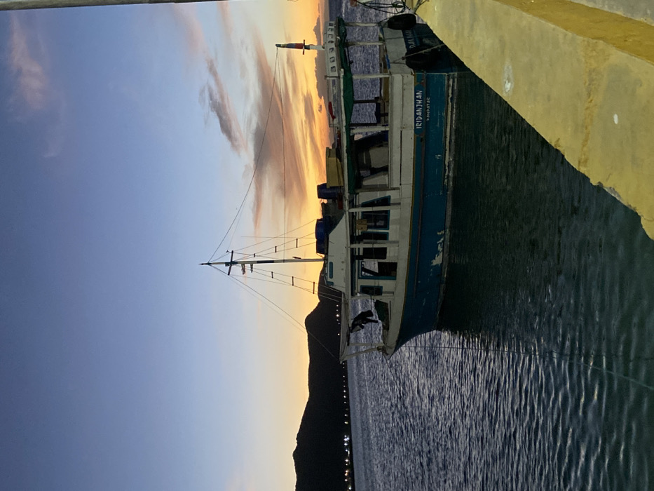 A fishing boat, "IRIDANJYAN," is docked beside a yellow concrete barrier. The vessel has a white upper deck and a blue hull. It is moored on rippling water. A dark silhouette of a mountain range and distant city lights are visible in the background. The sky transitions from blue to orange in the distance, creating a sunset or sunrise effect with the presence of light clouds.