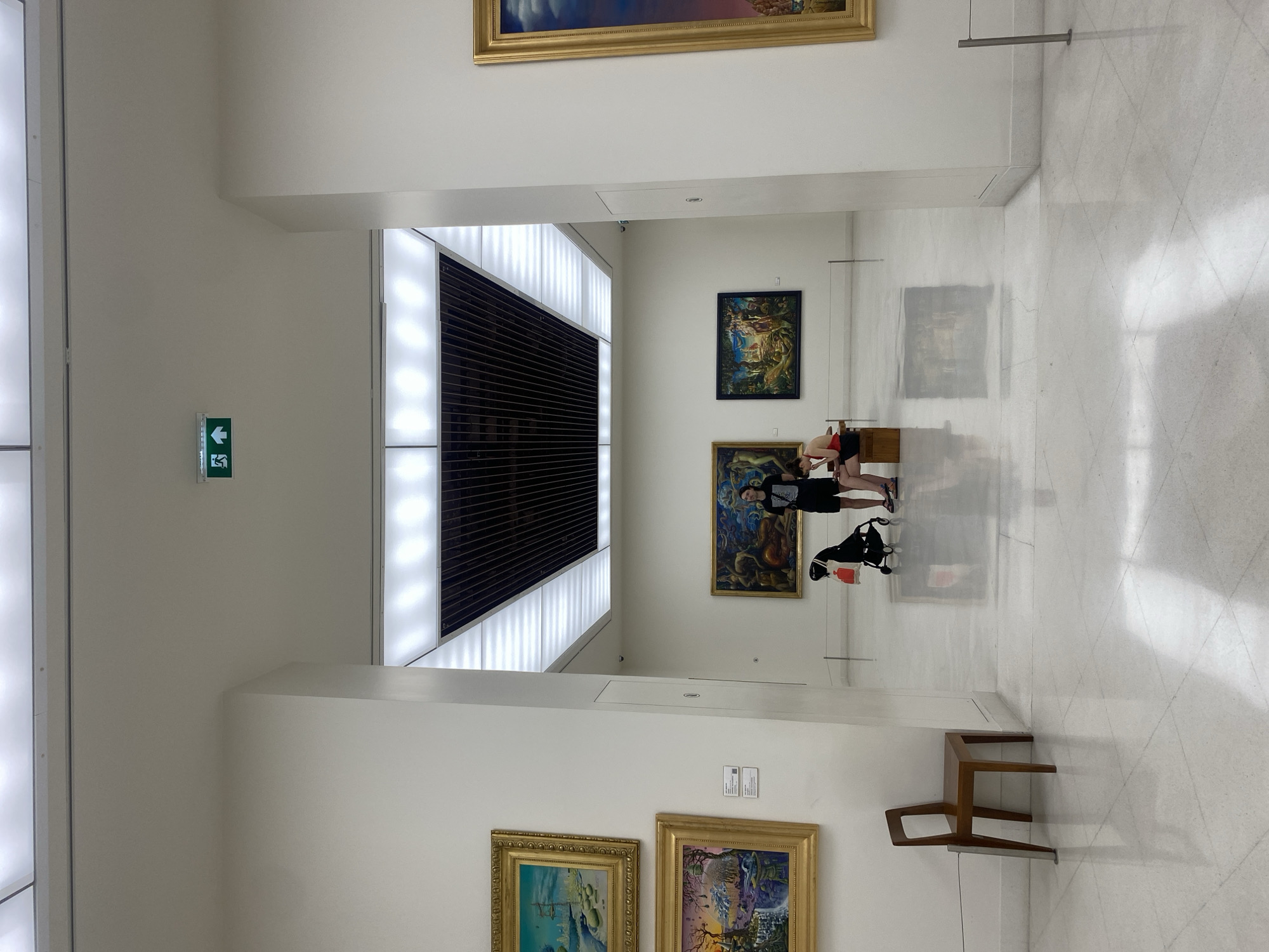 The image shows the interior of an art museum in Bangkok, Thailand. The museum has white walls and a white tiled floor. There is a long ceiling light fixture that runs down the center of the room, and art hangs on the walls. A woman wearing a black shirt and black shorts is visible in the middle of the room, kneeling down to look at an art display. A baby stroller is beside the woman. There is a chair in the foreground. An exit sign is on the left wall.