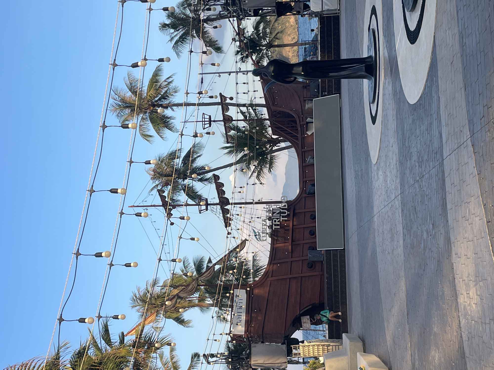 The image depicts a scene in Bac Nha Trang Ward, Vietnam. A decorative pirate ship facade stands amidst palm trees and a grid of strung lights. A bronze-colored statue is on the right side. A gray stone wall extends out from the right side of the ship facade. One individual is sitting at the base of the ship facade.