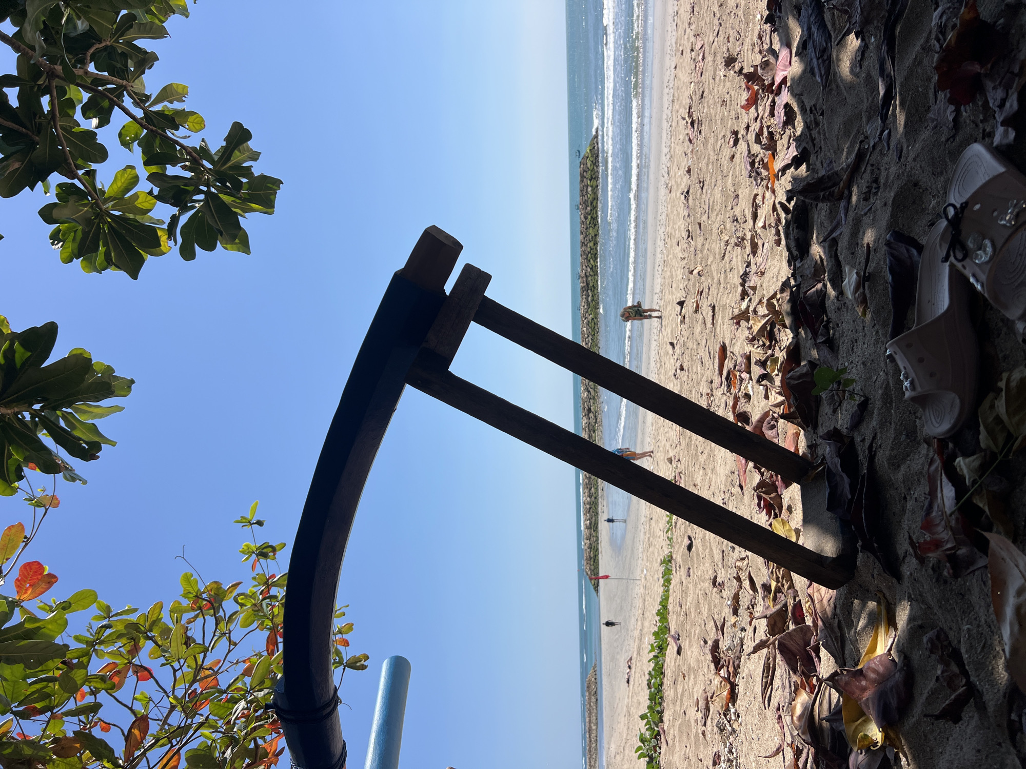 This is a low-angle, eye-level shot of a beach in Kuta, Indonesia. In the foreground, a pair of light brown flip-flops rests on the sand amongst fallen leaves. To the left, a dark curved structure, possibly part of a beach umbrella or structure, extends into the frame, with a hint of a light blue pole beneath it. Behind this, two dark wooden poles angled upwards suggest a support structure for something overhead. The middle ground features a sandy beach with a few scattered leaves and some green vegetation. In the background, the ocean meets the shore with gentle waves. Two stone breakwaters extend into the sea. Several people are visible on the beach, appearing as small figures in the distance, walking or standing. A red flag stands on a pole on the beach. The sky is a clear, bright blue. The leaves on the branches in the upper left of the frame display a mix of green and reddish-orange hues.