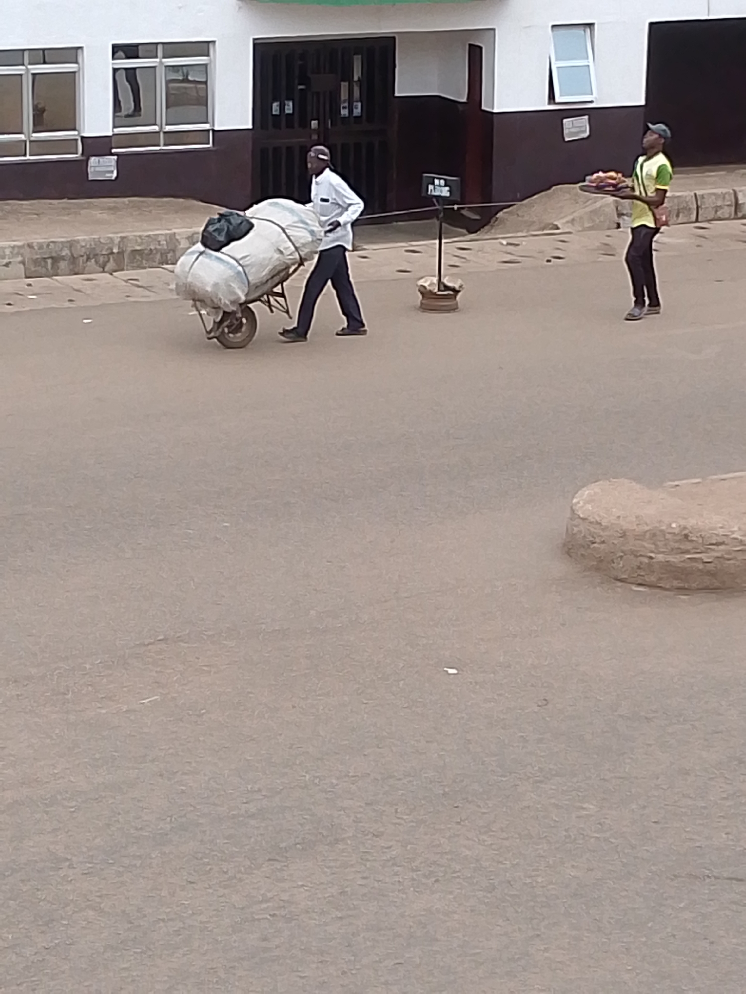 The image shows a street scene in Bukuru, Nigeria. One man is pushing a wheelbarrow loaded with a large bundle covered in white material with a dark bag on top. Another man stands nearby carrying a tray, possibly of food. The background includes a building with white walls, dark brown trim, and multiple windows and doors. There is also a sign that says "NO FLEEING". The road surface is a light brown color.