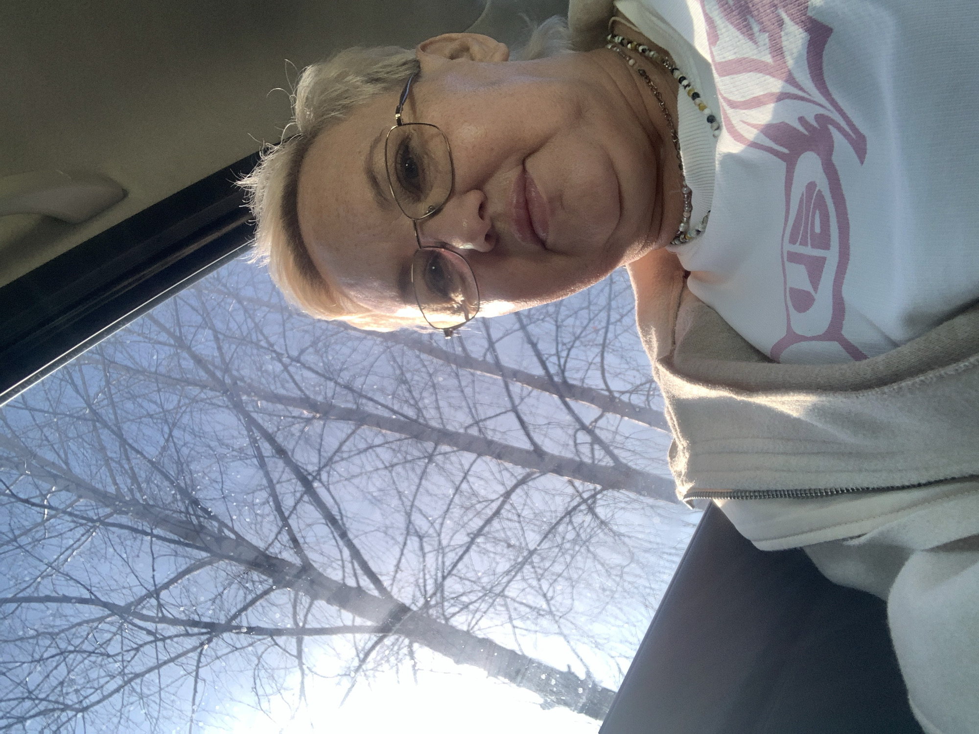 A selfie image showing an elderly woman with short blonde hair and glasses. She is wearing a white sweater with a light pink design and a beaded necklace. Behind her, viewed through a window, are bare tree branches against a bright blue sky. The location is presumed to be within the city of Chaikovsky, Russia.