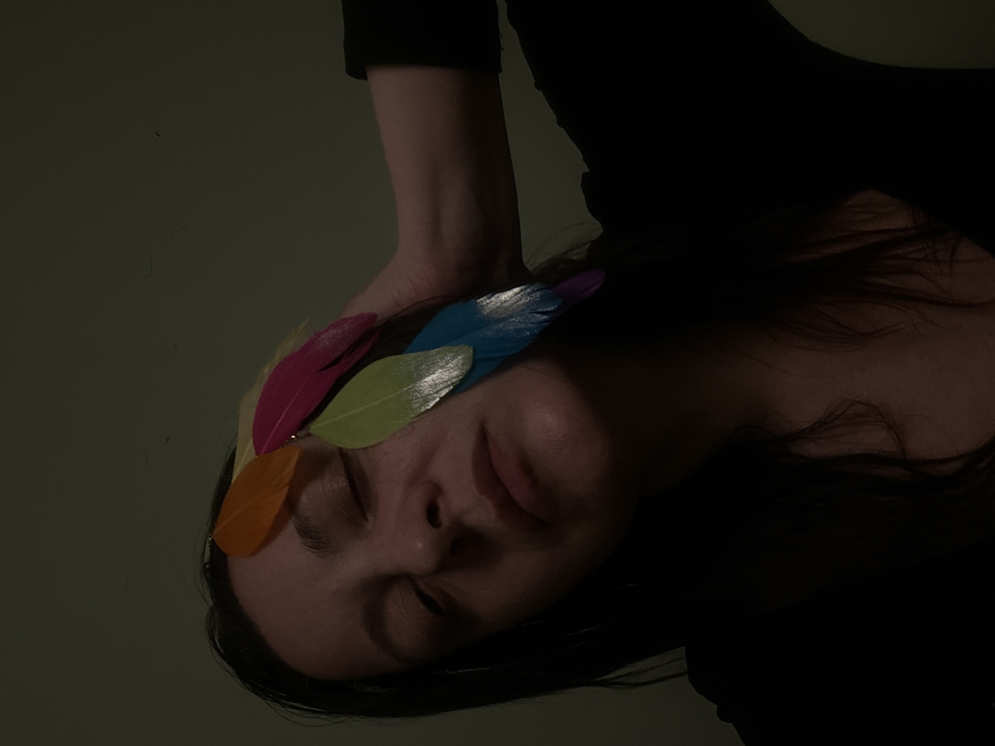 The media file presents a close-up, rotated portrait of a person, likely female, in a dim, indoor setting. The individual is shown lying down, with their head positioned on the right side of the frame. Their eyes are closed, and their facial expression appears serene or restful.
A cluster of vibrant, colorful feathers adorns their forehead and extends over their closed eyes. The feathers are in shades of hot pink, bright orange, lime green, light blue, and purple, some exhibiting a slight metallic sheen. These colorful elements provide a striking contrast against the muted tones of the person's skin and the dark background.
The person wears a dark, long-sleeved garment, parts of which are visible on their arm and upper body, blending into the deep shadows. Their dark hair cascades around their head, with some strands visible in the low light.
The background is a plain, dark greenish-gray surface, suggesting a simple wall or backdrop. The lighting is low-key, creating soft shadows and highlighting the contours of the face and the texture of the feathers, contributing to a tranquil and slightly enigmatic atmosphere. There is no visible text in the image.