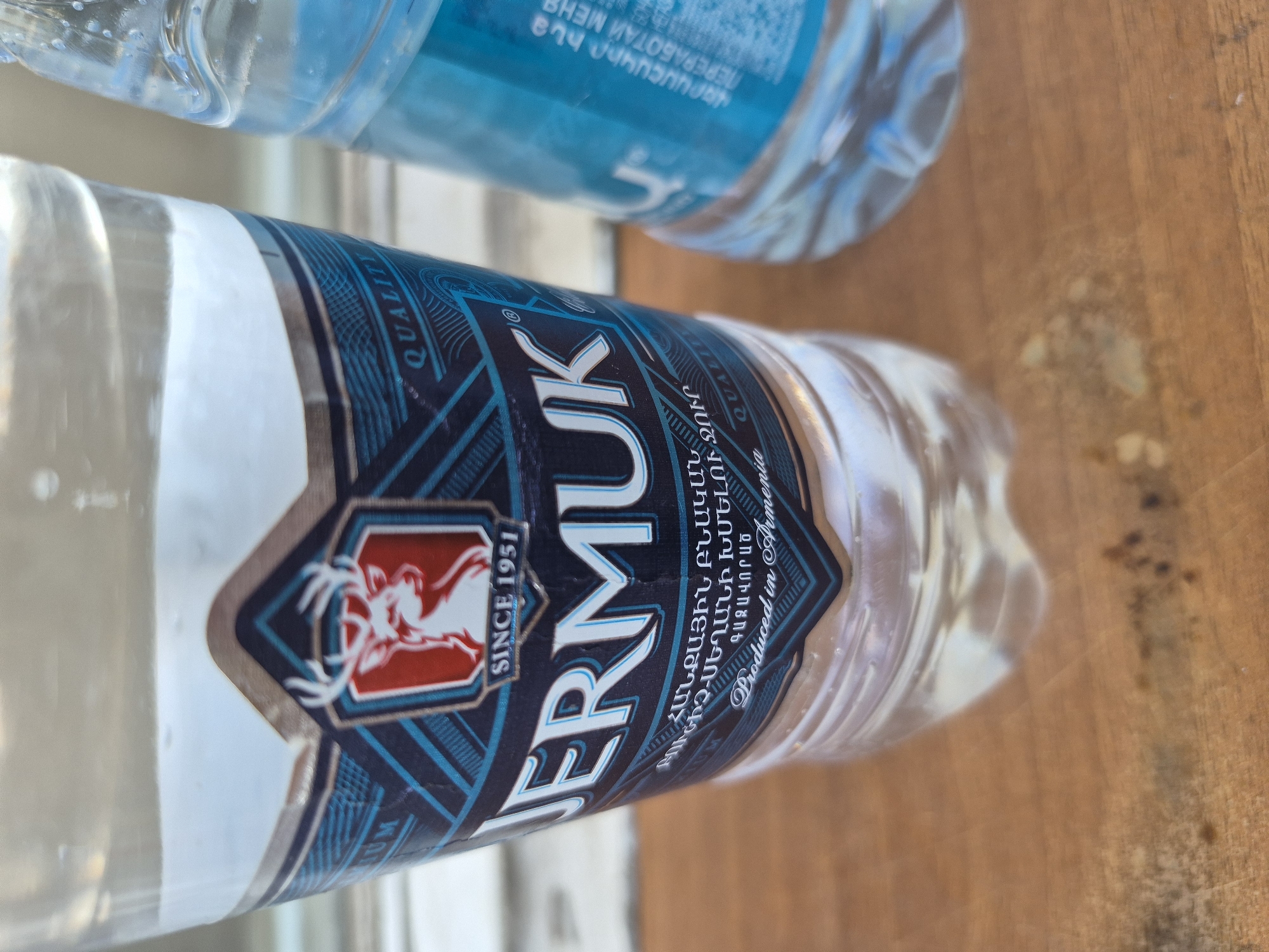 The image displays two plastic water bottles on a light brown wooden surface, likely a table or countertop, illuminated by bright daylight. The primary subject is a clear water bottle positioned diagonally, prominently featuring a dark blue, red, and white label. The brand name "JERMUK" is clearly visible in white capital letters. The label also displays a red emblem with a white, antlered creature, the text "SINCE 1951," and "QUALITY." Below the brand name, Armenian text states "ՀԱՆՔԱՅԻՆ ԲՆԱԿԱՆ" (Mineral natural), "ԲՈՒԺԻՉ-ՍԵՂԱՆԻ ԽՄԵԼՈՒ ՋՈՒՐ" (Medicinal-Table Drinking Water), and "ԳԱԶԱՎՈՐԱԾ" (Carbonated). The English phrase "Produced in Armenia" confirms the origin, aligning with the Yerevan, Armenia context.
In the upper right background, another plastic water bottle is partially visible and out of focus. This bottle has a light blue label with mirrored text, including "ПЕРЕРАБОТАЙ МЕНЬ" and "ՎԵՐԱՄՇԱԿԻՐ հա" (Recycle me in Russian and Armenian, respectively). No people or specific events are depicted, making the bottles the central focus of the composition.