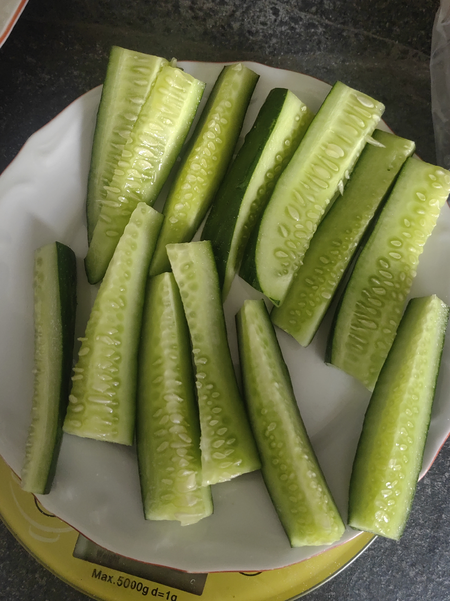 A plate of sliced cucumbers is shown, resting on a yellow digital scale. The cucumbers are bright green and cut lengthwise into approximately 1-inch wide strips. Some pieces are whole strips, while others are halved lengthwise. The seeds and white flesh of the cucumber are visible in the cut surfaces. The plate is white with a red rim and is positioned on the scale. The background appears to be a dark, textured surface, possibly a countertop. The scale displays "Max. 5000g d=1g" in black text. The scene is indoors and appears to be in a kitchen. There is no information in the image that indicates the location is in the city of Djerovo, Bulgaria.