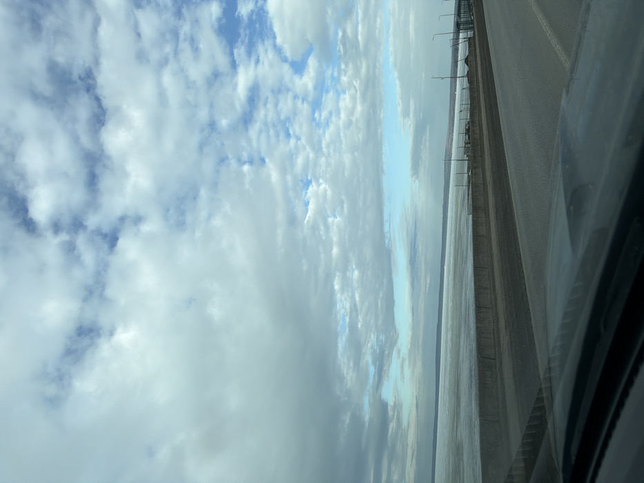 This image captures an outdoor scene in Chaikovsky, Russia, looking across a partially frozen body of water, likely the Votkinsk Reservoir, from a viewpoint inside a vehicle. The sky occupies the upper two-thirds of the frame, filled with a mix of stratocumulus and cumulus clouds, primarily in shades of gray and white, with distinct patches of blue sky visible between them. The middle ground features a wide expanse of water, predominantly covered with sheets of ice or slush, giving it a muted, reflective appearance. A low, dark landmass is visible on the distant horizon, bordering the water. The foreground shows the upper edge of a vehicle's dashboard, dark in color, reflecting the sky. Immediately beyond the vehicle interior is a multi-lane asphalt roadway or bridge surface. A dark, robust concrete barrier runs parallel to the roadway, separating it from the icy water. To the right side of the frame, past the barrier, a strip of land or another section of the bridge features several dark, vertical poles, presumably streetlights, and some fencing or structural elements extending into the distance. No individuals, specific actions, or interactions are discernible.