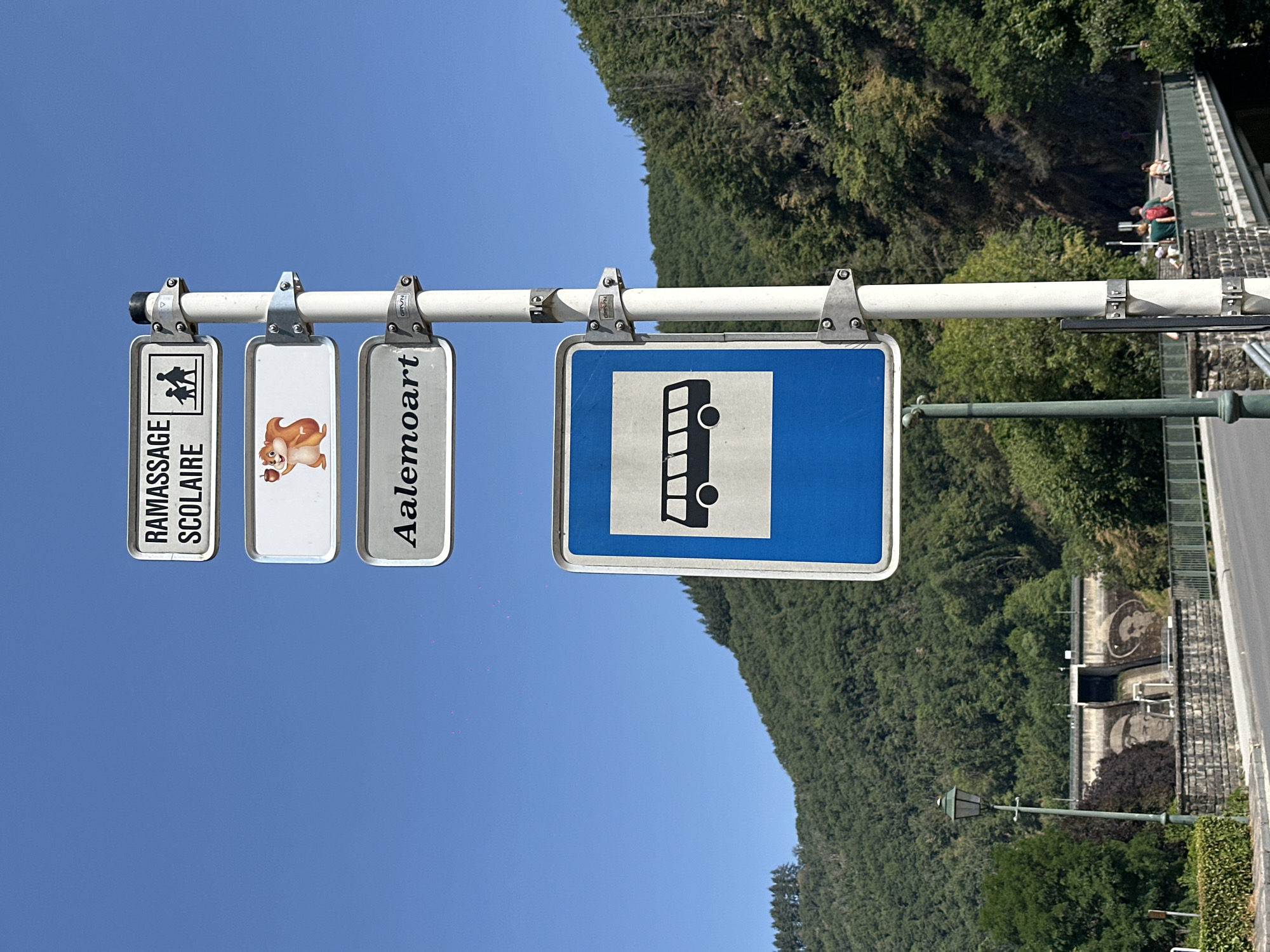 The image captures a street scene in Vianden, Luxembourg, focusing on a pole with multiple signs and a bus stop. The pole, made of metal, is positioned on the left side of the frame. Attached to it are four signs. The top sign, rectangular with a white background and black text, reads "RAMASSAGE SCOLAIRE" and features a pictogram of two children walking, indicating a school pick-up point. Below this is a rectangular sign with a white background and a cartoon image of a squirrel holding an acorn. The third sign, also rectangular with a white background and black text, displays "Aalemoart". The lowest sign is a blue rectangle with a white border, depicting a black silhouette of a bus, signifying a bus stop.
In the background, a lush green forested hill rises, and at its base, a dam is visible with two large painted faces on its structure. A lamppost stands to the left of the signs, and a bridge with a green railing spans across a body of water or a road. Several people are seen crossing the bridge. The sky is a clear, bright blue.