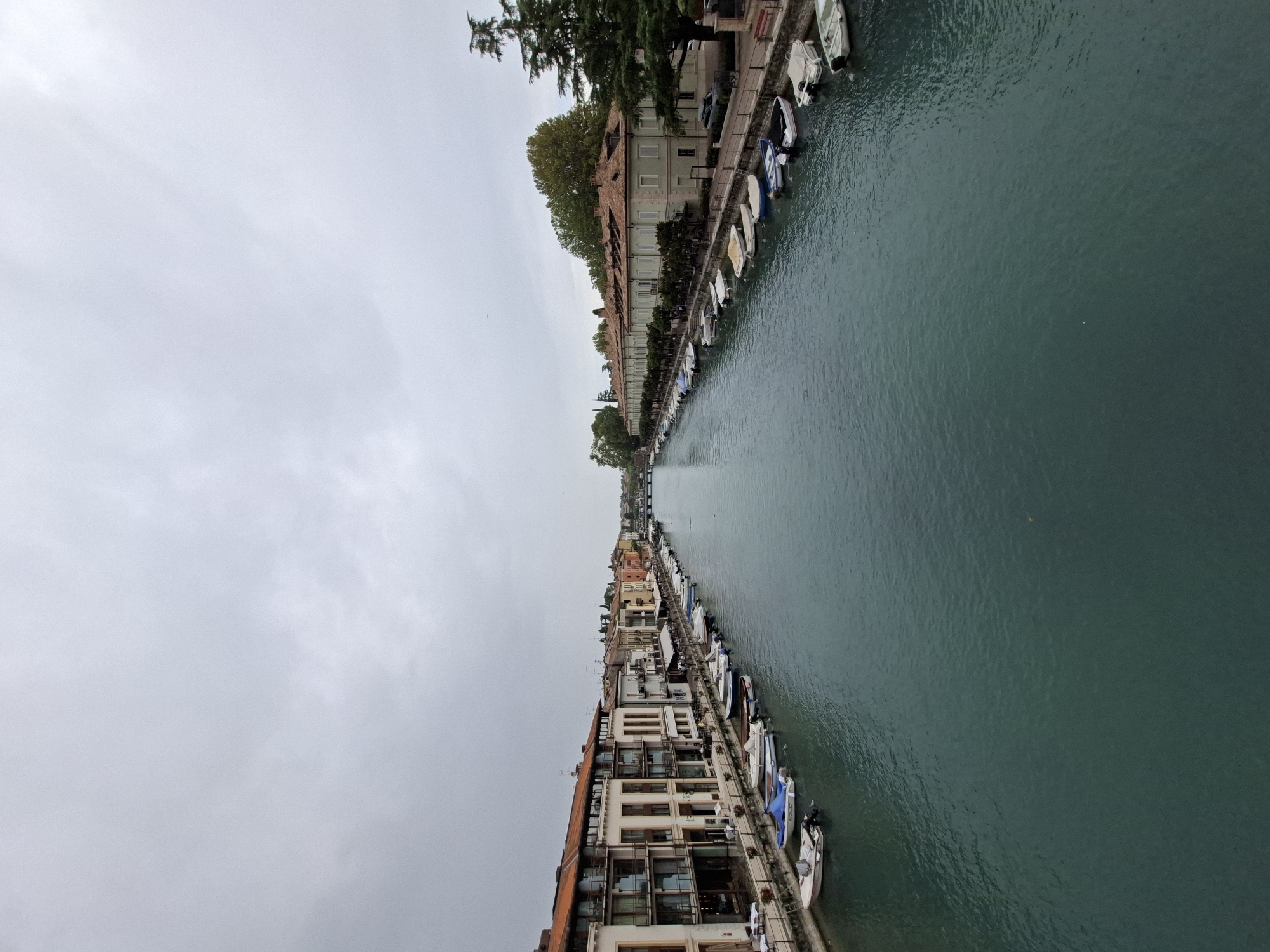 The image shows a view of a canal in Peschiera del Garda, Italy, on an overcast day. The canal is lined with buildings on both sides, featuring residential and commercial properties with terracotta roofs. Numerous small boats are moored along the edges of the canal, some covered and some uncovered. A pedestrian walkway is visible alongside the buildings. The water of the canal is a deep turquoise color, with ripples indicating a slight current or breeze. In the distance, a bridge can be seen spanning the canal, and trees and greenery dot the landscape beyond the buildings. The sky is uniformly gray, suggesting cloudy weather.