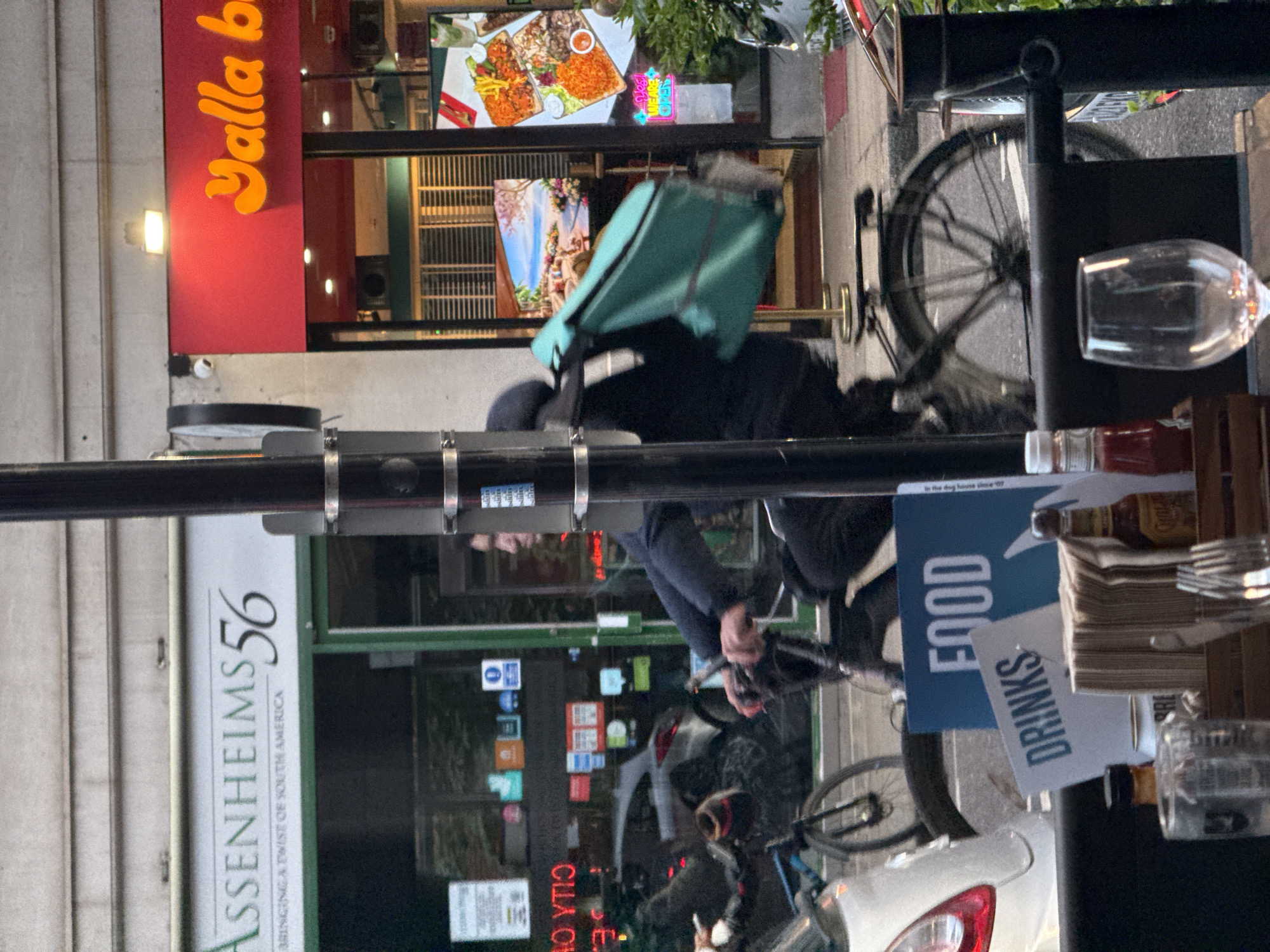 Here is a description of the image:
The image, taken at street level, features a man riding a bicycle with a delivery backpack in front of a restaurant. The restaurant on the right is "Yalla b", while on the left, the other establishment is named "Assenheims56". In the bottom right of the frame is a sign promoting "Drinks" and "Food".