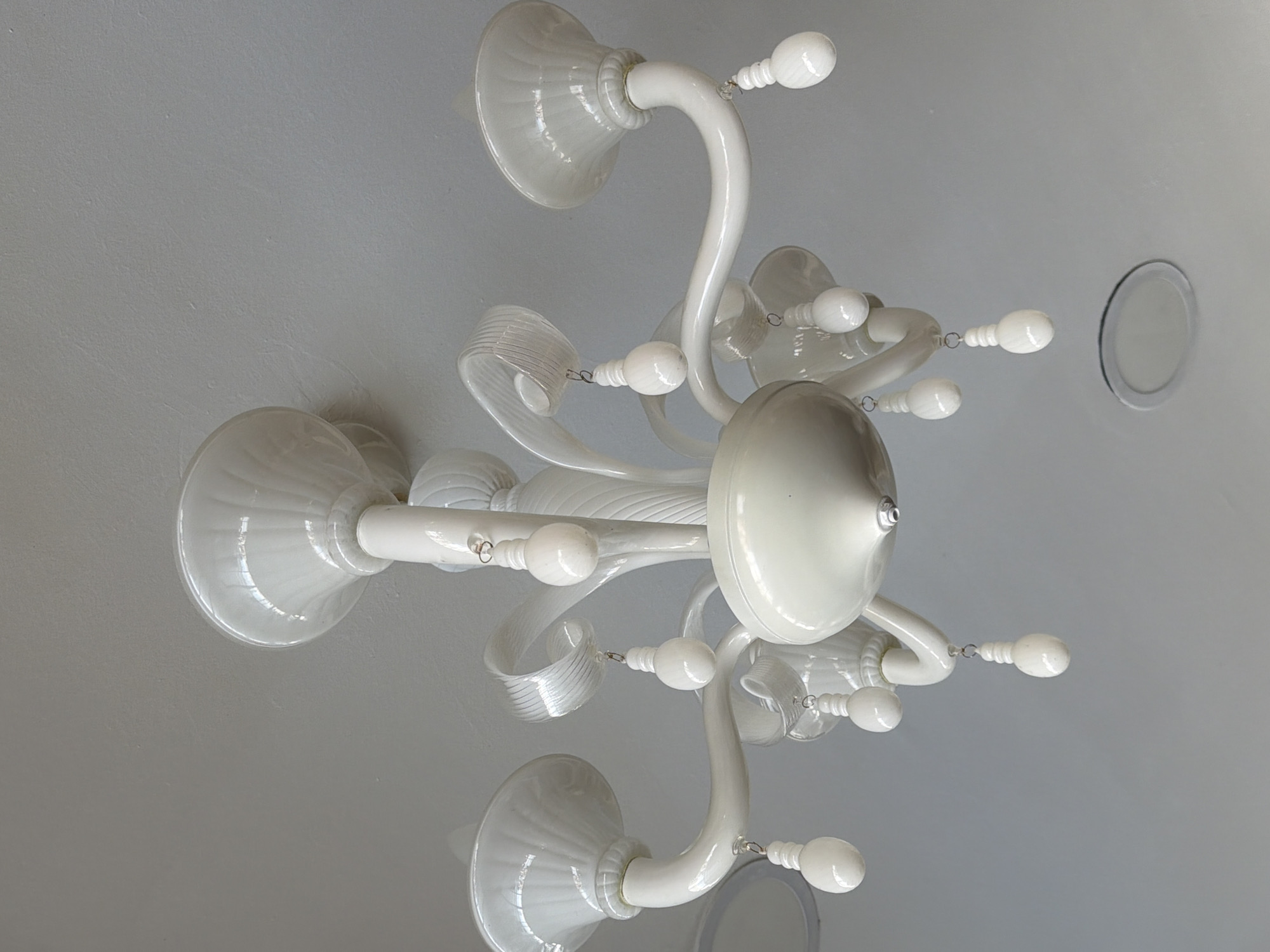 A white, ornate chandelier hangs from a plain white ceiling. The chandelier has multiple arms, each supporting a light fixture. Some of the fixtures are bell-shaped, while others are more decorative, with ribbed glass accents. Small, teardrop-shaped white ornaments hang from chains attached to the arms of the chandelier. The overall aesthetic is classic and somewhat elaborate. The scene is illuminated by natural light, suggesting it is daytime. There are no people or animals visible in the image. The ceiling has a subtle texture and there is a circular recessed light fixture visible in the lower right portion of the image. The location is identified as Marmaris, Turkey.