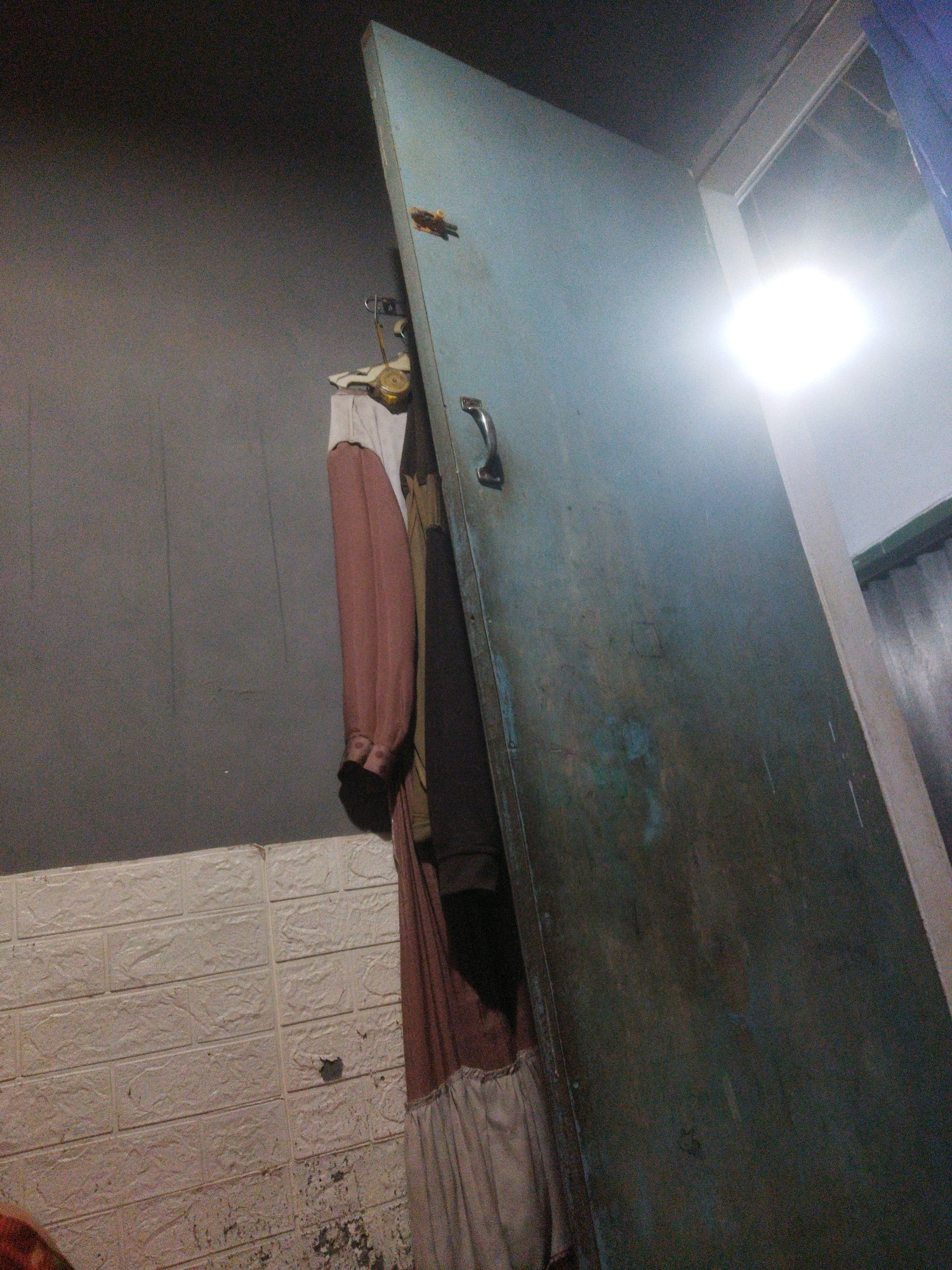 The image depicts a dimly lit interior space, likely a residential room in Coban Joyo, Indonesia. A large, weathered, blue-green metal door is prominently positioned, slightly ajar and leaning against a wall. The door has a silver handle. To the left of the door, several articles of clothing hang from a rack, including a long pink garment with a white trim and another dark garment. The wall behind the clothing is a textured gray. In the lower left portion of the image, a white tiled wall with a brick pattern is visible, showing signs of wear and tear. A bright light source emanates from above and to the right, suggesting an open doorway or a window. The overall impression is one of a utilitarian and somewhat neglected space.