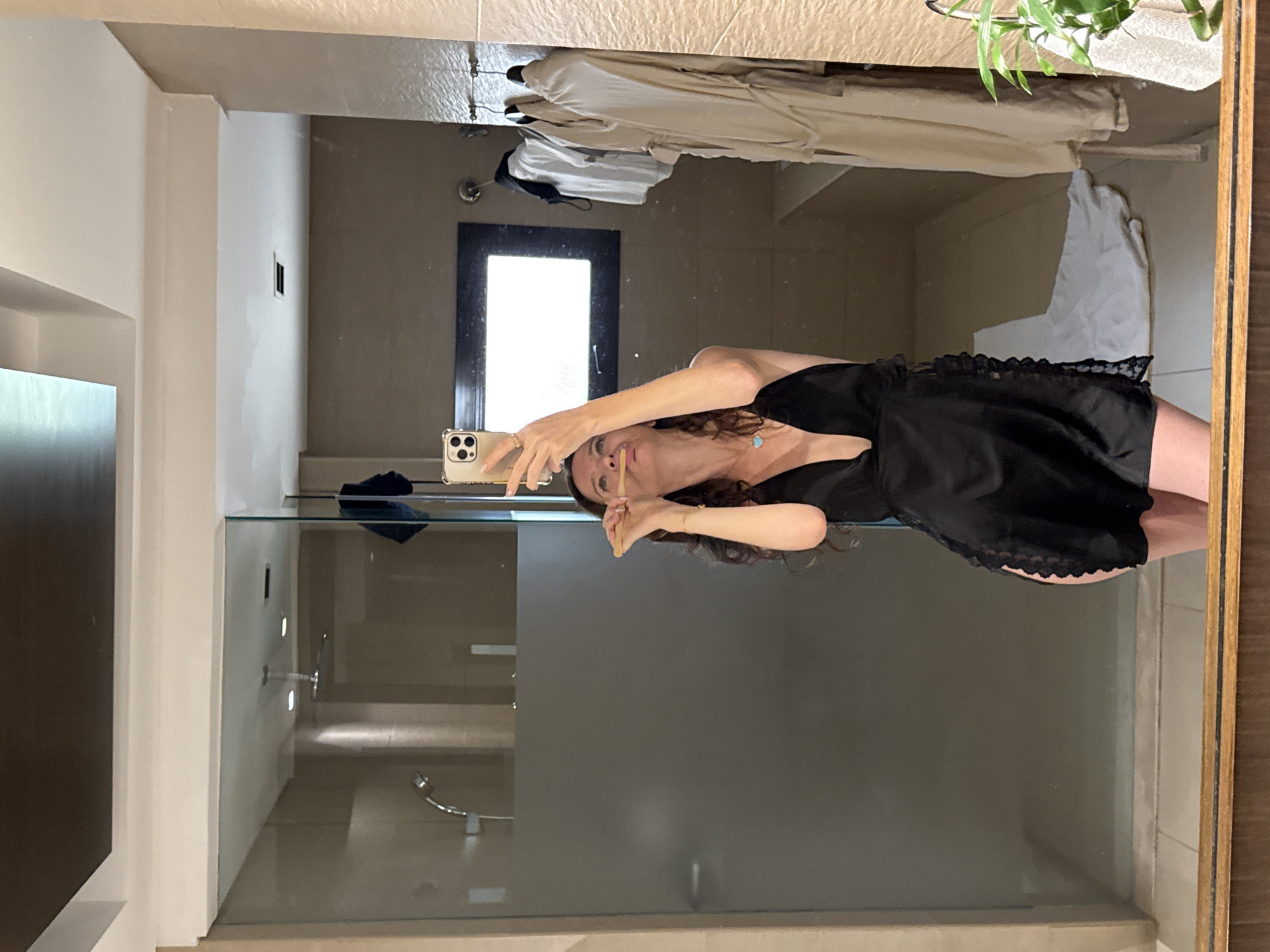 A woman is taking a mirror selfie in a modern bathroom, holding a wooden toothbrush to her mouth. She is wearing a black silk slip dress with lace trim and a light blue pendant necklace. In the background, a glass shower door is visible, with a showerhead inside. On a rack to the right, several light-colored robes are hanging. The bathroom appears to be in a hotel or upscale residence. The scene is likely in Dubai, United Arab Emirates, given the context provided by the prompt.