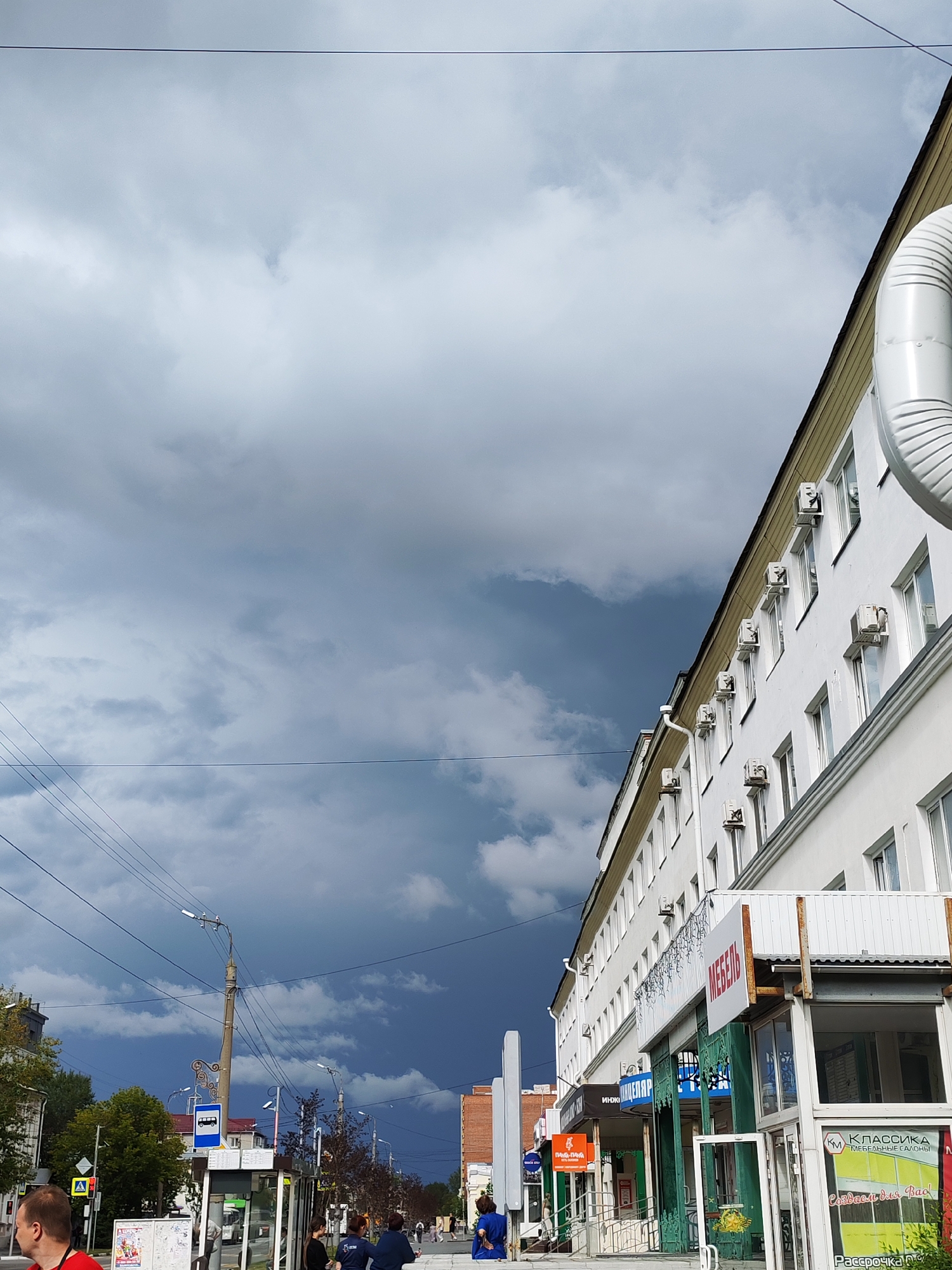 The image depicts an outdoor scene in an urban environment, likely a street. The sky is overcast with dramatic, dark clouds dominating the upper portion of the frame, suggesting impending rain or a storm. In the foreground and midground, a white building with multiple windows and air conditioning units lines the street. There are several storefronts visible on the ground floor of this building, with various signs, including one that reads "МЕБЕЛЬ" (Furniture) in red lettering.
To the left of the building, a bus stop shelter with a sign indicating bus routes is visible. Several utility poles with wires stretch across the scene. Trees with green and some autumn-colored foliage are present, particularly on the left side of the image.
There are a few individuals scattered throughout the scene. In the far left foreground, a person wearing a red shirt is partially visible. Further down the street, near the bus stop and storefronts, several people are seen standing or walking. One person in a blue jacket appears to be interacting with another person. In the midground, more figures are walking along the sidewalk.
The overall impression is a city street under a heavy, stormy sky, with commercial activity and pedestrian presence. The location is identified as Unknown, Unknown, with no specific geographical markers.