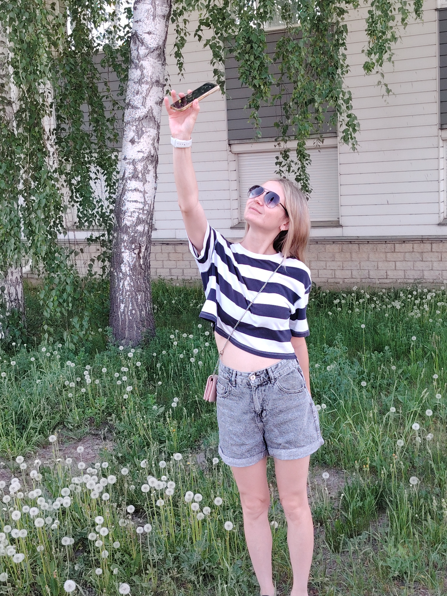 A woman stands in a field of dandelions in front of a birch tree in Chaikovsky, Russia. She is wearing a striped crop top, denim shorts, and sunglasses. She is holding a smartphone up in the air, possibly taking a photograph or video. Behind her is a light-colored building with gray trim and a closed shuttered window. The sky is overcast.