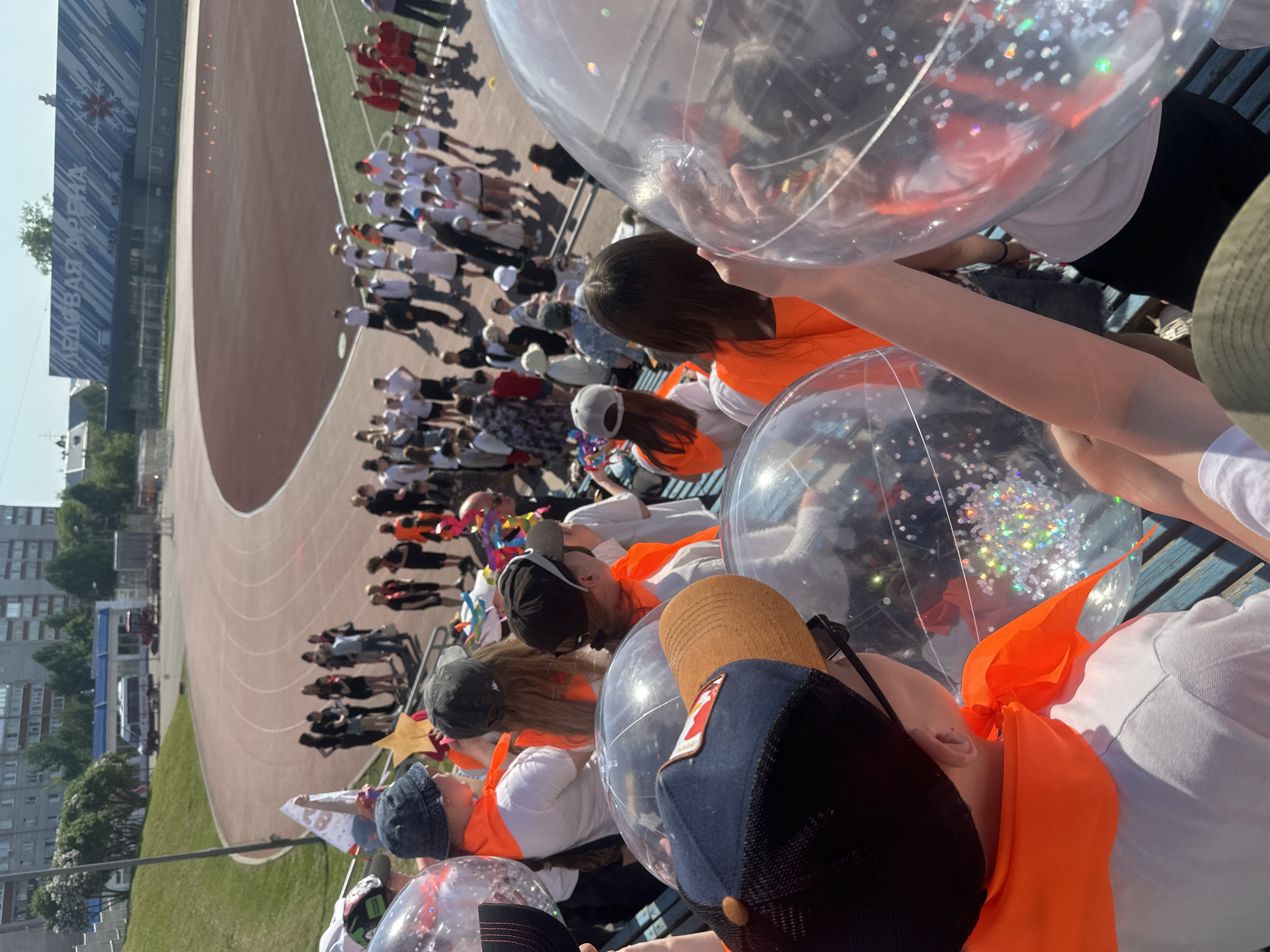 People sit on benches in a stadium, holding clear balloons with confetti. Many wear orange vests. A track surrounds a green field. In the background, more people stand on the field. Buildings and a clear sky are visible beyond the stadium. The location is Votkinsk, Russia, as indicated by the building in the background labeled "Ice Palace Votkinsk Arena" in Russian.