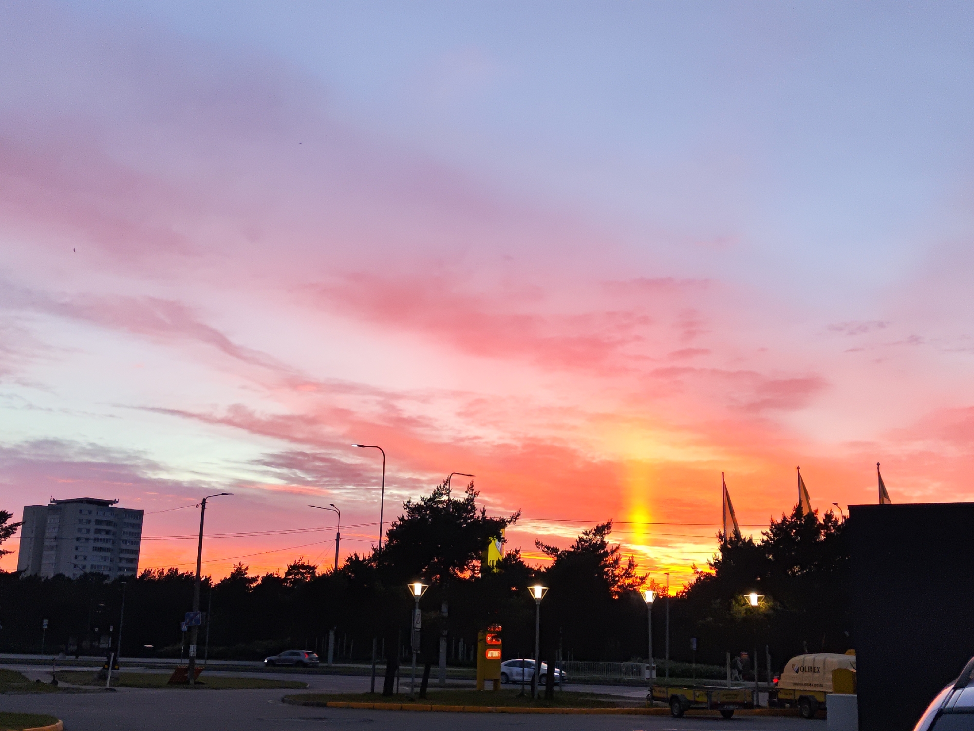 The image captures a vibrant sunset over a city, likely Tallinn, Estonia, given the distinctive architecture of the building and the presence of trees commonly found in the region. The sky is a dramatic display of color, transitioning from soft blues and purples at the top to vivid pinks and fiery oranges near the horizon. A distinct vertical shaft of yellow light pierces the sky, a phenomenon known as a sun pillar, caused by light reflecting off ice crystals in the atmosphere.
In the foreground, a street intersection is visible, with asphalt roads and a grassy median. Several streetlights line the road, some illuminated, casting a warm glow. Parked on the side of the road are two trailers, one with the brand name "OLIREX" visible. A dark, modern building partially obscures the right side of the frame.
Behind the street, a dense line of dark trees forms a backdrop. To the left, a tall, gray, multi-story residential building stands prominently against the colorful sky. A few cars are present on the road, including a silver sedan and another partially visible vehicle. In the distance, three yellow flags with dark symbols are seen flying above the trees. The overall scene suggests an urban environment at dusk, with the natural beauty of the sunset being the focal point.