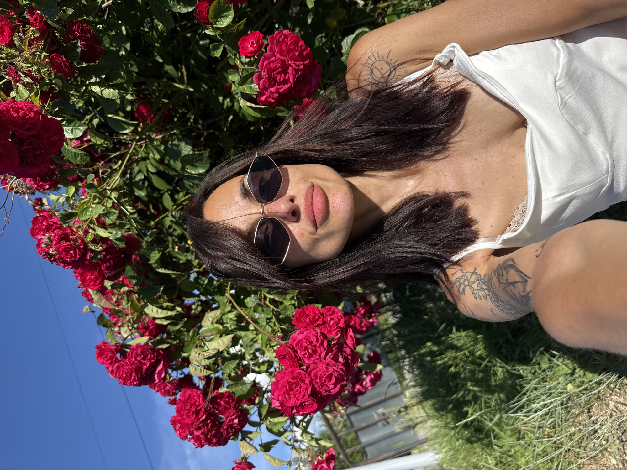 In a bright, outdoor setting possibly in Lovech, Bulgaria, a young woman with dark hair and sunglasses is captured in a close-up selfie. She is positioned in front of a vibrant rose bush laden with deep red blossoms. The woman wears a white camisole with delicate lace trim. Her left arm, visible in the foreground, displays a tattoo. The background features lush green grass and a clear blue sky, with a hint of a fence and some utility lines. The sunlight casts a warm glow on the scene, highlighting the textures of the flowers and the woman's features.