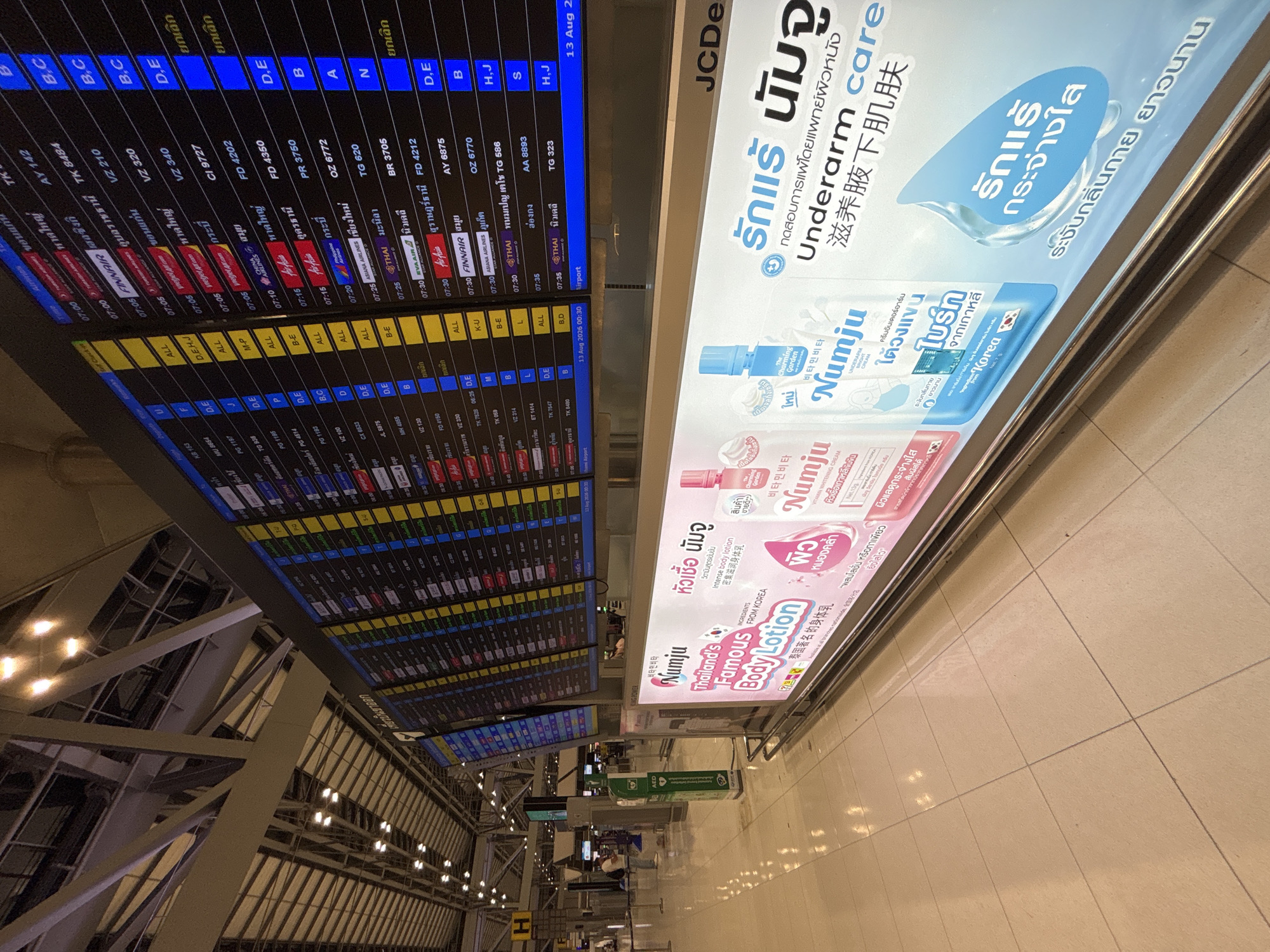 The image shows an interior view of an airport terminal in Nong Prue Subdistrict, Thailand. The primary focus is on a large departure board displaying flight information and a prominent advertisement for a body lotion product. The departure board shows multiple rows of flights with details such as airline, destination, flight number, and departure time, with some information displayed in Thai script. The advertisement features the brand "Numju" and highlights "Thailand's Famous Body Lotion" and "Underarm care," also with Thai text. In the background, people are visible walking and waiting. The environment appears to be a modern airport concourse with polished flooring and overhead lighting.