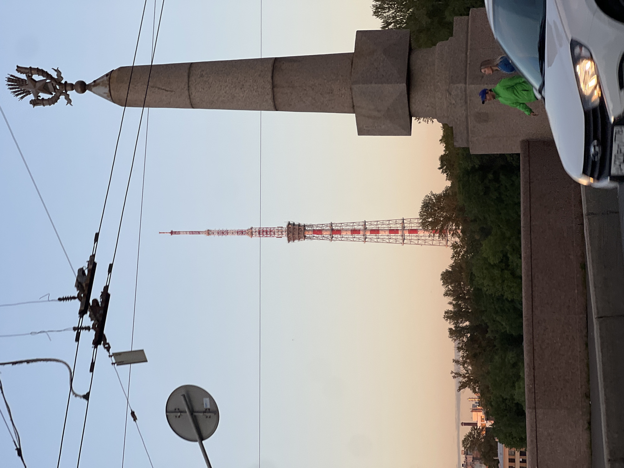 The image captures a view of the Saint Petersburg Television Tower and the Rostral Column on Vasilievsky Island, Russia, during what appears to be sunset or early evening. In the foreground, the top of a white car is visible. To the right, a large stone obelisk, topped with a decorative emblem featuring a star and laurel wreath, stands prominently. Two individuals, a man in a green jacket and blue cap and a woman in a blue top, are walking past the obelisk. Behind them, a dense line of trees obscures the lower part of the city skyline, with buildings peeking through the foliage. The Saint Petersburg Television Tower, a tall, red-and-white lattice structure, dominates the background, with overhead wires and a traffic signal pole visible in the upper left quadrant. The sky displays a gradient of warm, soft colors typical of sunset.