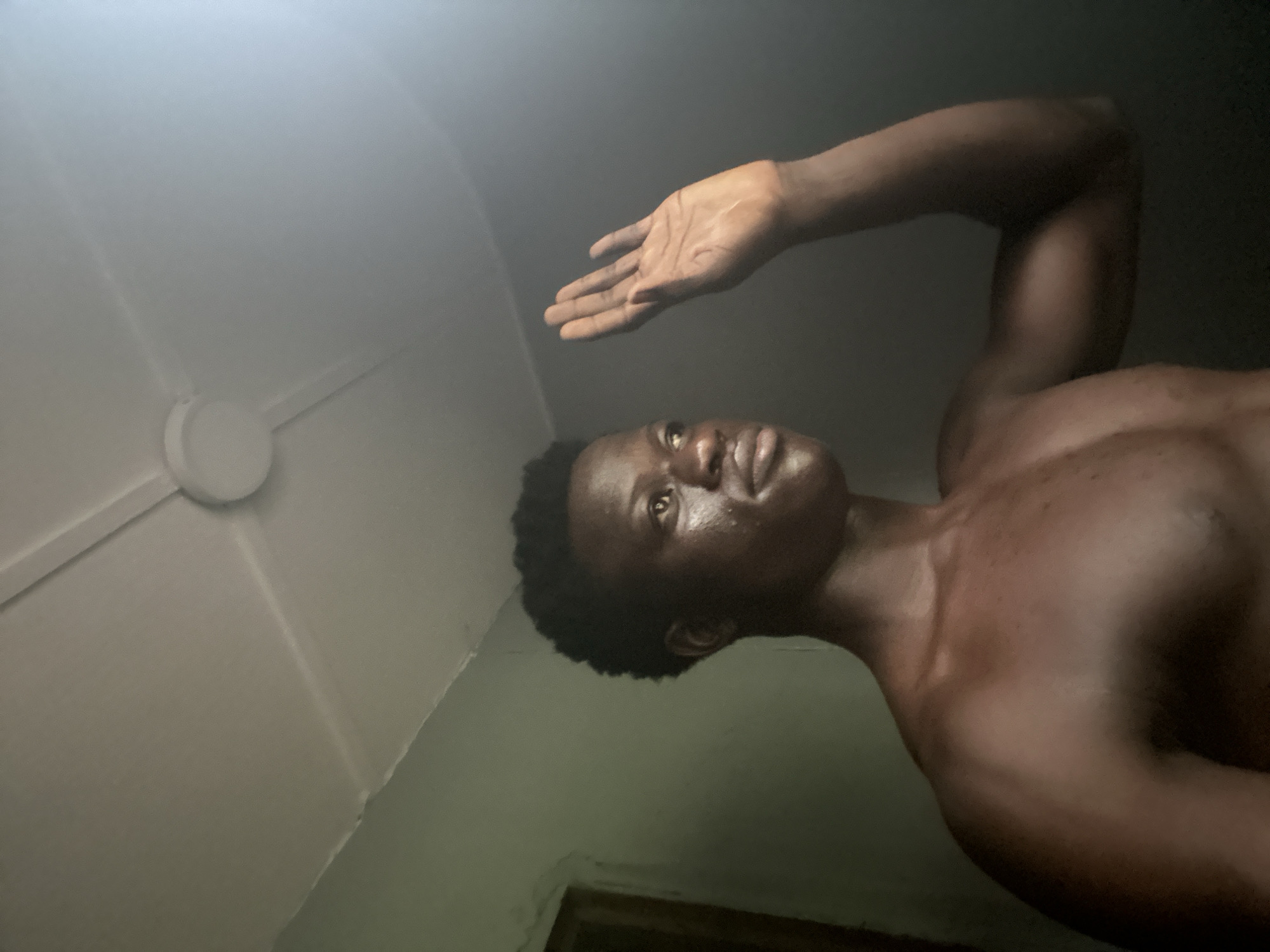 A young Black male is shown from the chest up, facing the left side of the frame. His skin is dark and appears slightly dewy. He has short, dark curly hair. His left arm is raised with his palm facing outwards, as if waving or warding something off. A faint light source is visible to the right of his face and arm, creating a bright glare in the upper right corner of the image. The background consists of a light-colored ceiling with visible seams and a ceiling fan. The walls appear to be a muted green color, with a dark doorway visible on the far left. The overall lighting is dim, with the primary light source coming from the right. The scene is consistent with an indoor setting, possibly a bedroom or living space, in the city of Badagry, Nigeria.