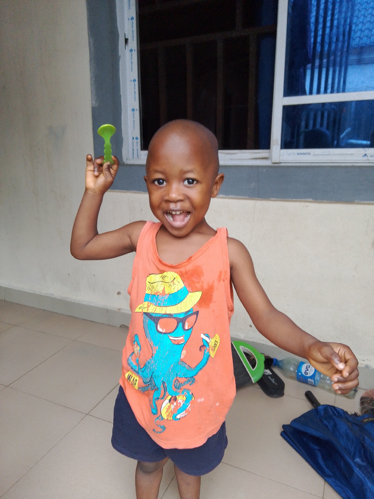A young boy stands outdoors on a tiled surface, holding a green toy in his raised right hand. He is wearing an orange sleeveless t-shirt with a graphic of a blue octopus wearing a hat and sunglasses, and dark blue shorts. The boy is looking directly at the camera with a wide, open-mouthed smile. Behind him, a wall with a window can be seen. To the right of the boy, on the tiled ground, is a blue umbrella, a plastic water bottle, and a green object which appears to be a dustpan. The environment suggests a residential or communal outdoor area.