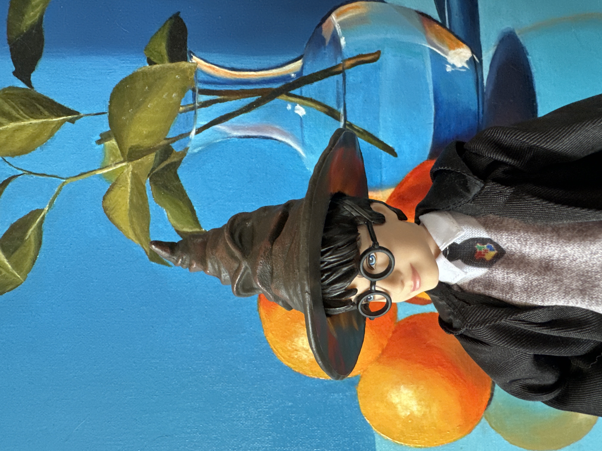 A close-up, vertical shot depicts a Harry Potter doll wearing a Sorting Hat and a black and gray uniform in front of a painting. The doll is positioned in the center of the frame, with its head tilted slightly to the left. The background painting features a blue backdrop with green leaves and branches on the left, a clear glass vase with visible water distortion in the upper center, and orange fruits scattered around the bottom. No specific details suggest the location is in Krasnoyarsk, Russia.