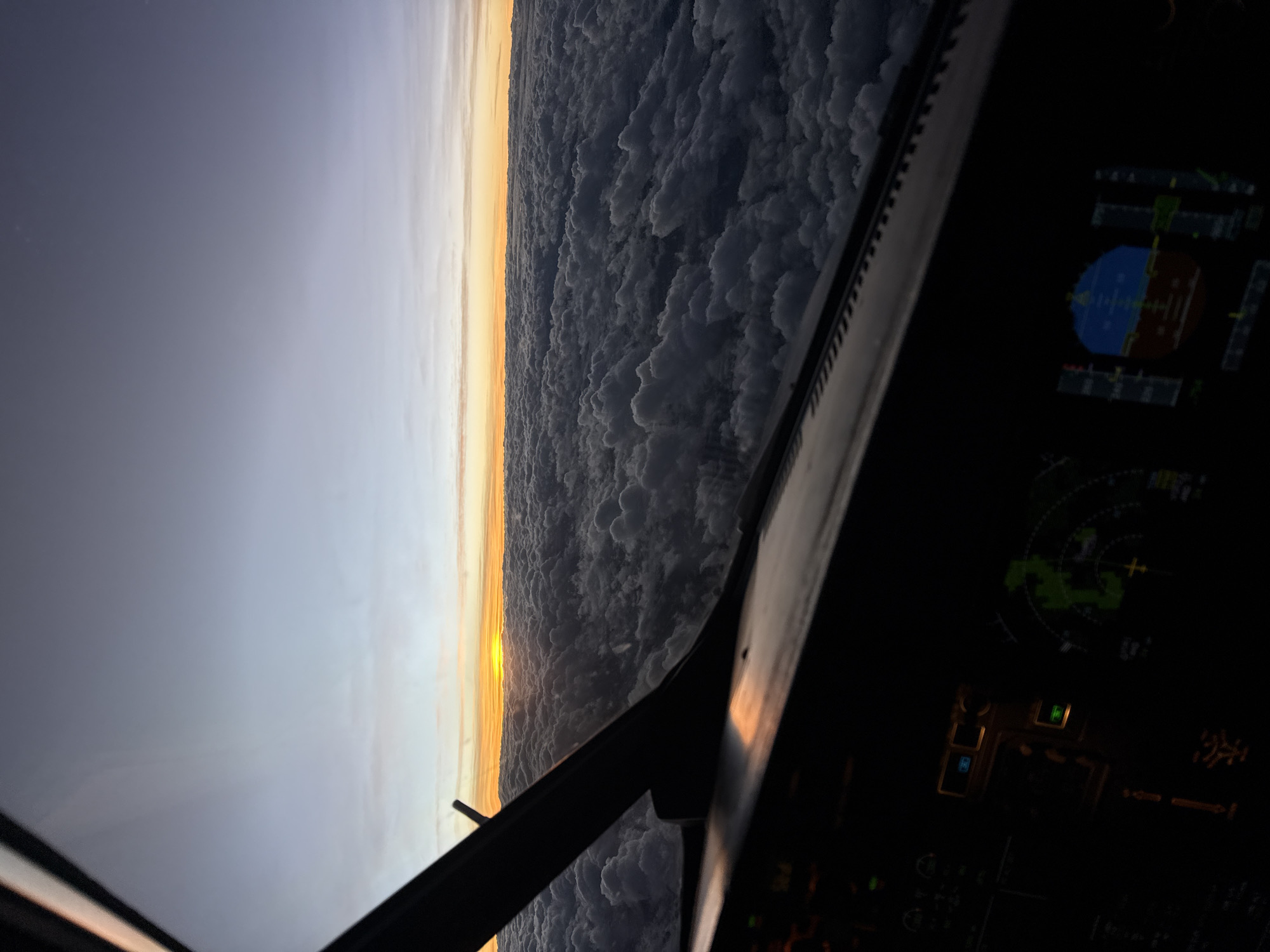 The image shows a view from inside an airplane cockpit. Outside, a sunrise or sunset is visible on the horizon, with a thick layer of clouds below. The cockpit instrumentation panel is partially visible and lit up. The location is not determinable from the image. There is no indication of the city of Tintay, Peru in the image.