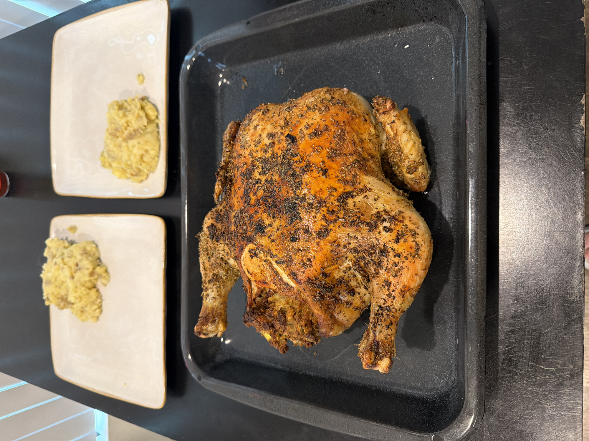 A freshly roasted whole chicken is presented on a black baking sheet, accompanied by two square plates, each containing a portion of yellow, mashed food. The setting appears to be a domestic kitchen, with a black countertop and visible kitchen elements. This scene is likely to be found in a home in Los Angeles, United States of America.