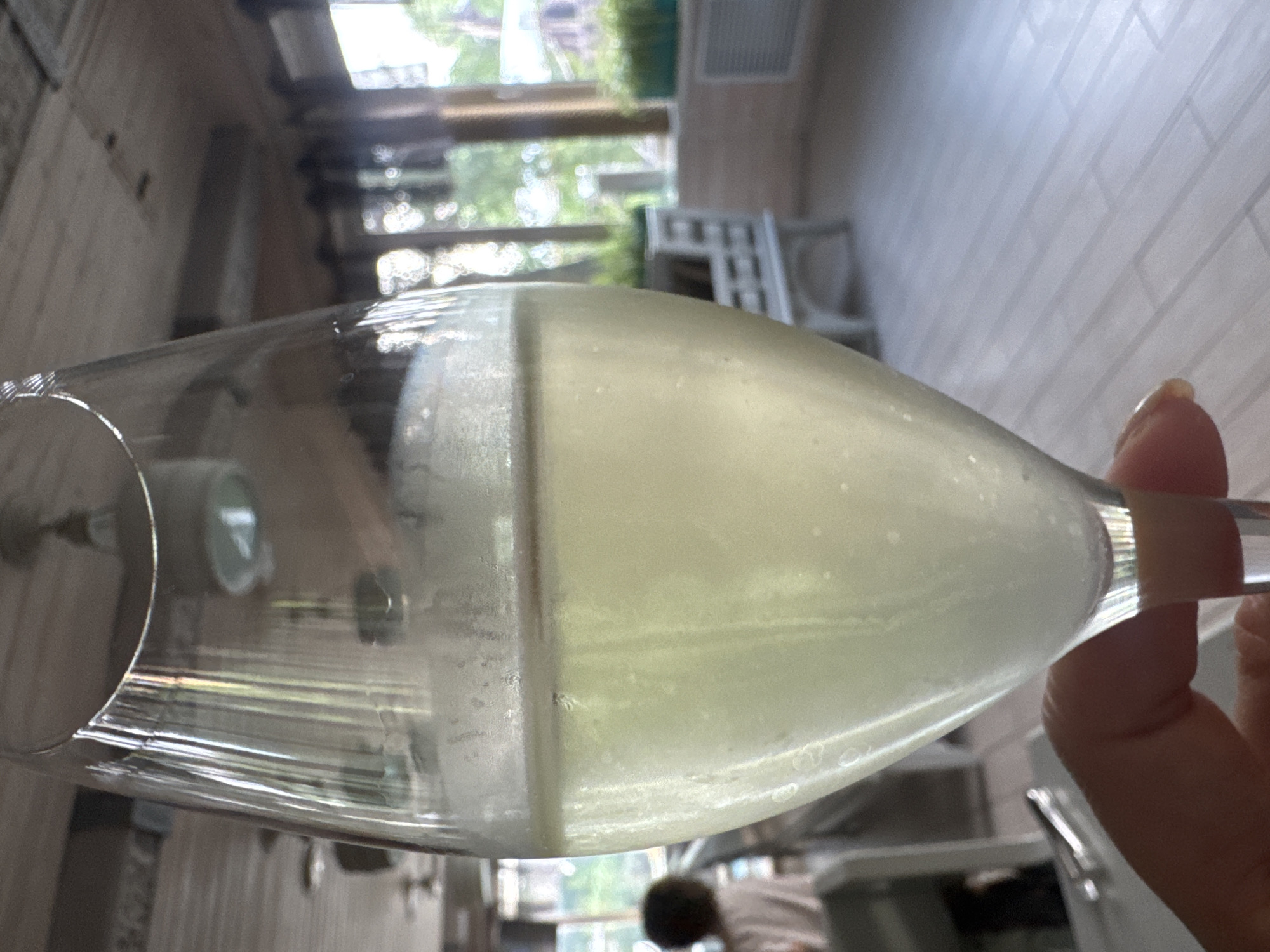 A hand holds a champagne flute filled with a pale yellow beverage, likely champagne or a similar sparkling wine, with visible bubbles and foam. The glass is condensation-covered, indicating it is cold. The background is blurred, suggesting an indoor or semi-outdoor setting. Elements visible in the background include wooden ceiling beams, light fixtures, and windows that reveal greenery and outdoor structures, possibly a restaurant or cafe. A person with dark, curly hair, wearing a light-colored shirt, is seated and out of focus in the middle ground to the left. To the right, a white bench-like structure and planters with green foliage are visible. The floor appears to be made of light-colored, wide planks. The overall impression is of a casual, possibly celebratory moment in a pleasant environment. This location is not identifiable as the city of Sovkhoza imeni Lenina, Russia, based solely on the visual information provided in the image.