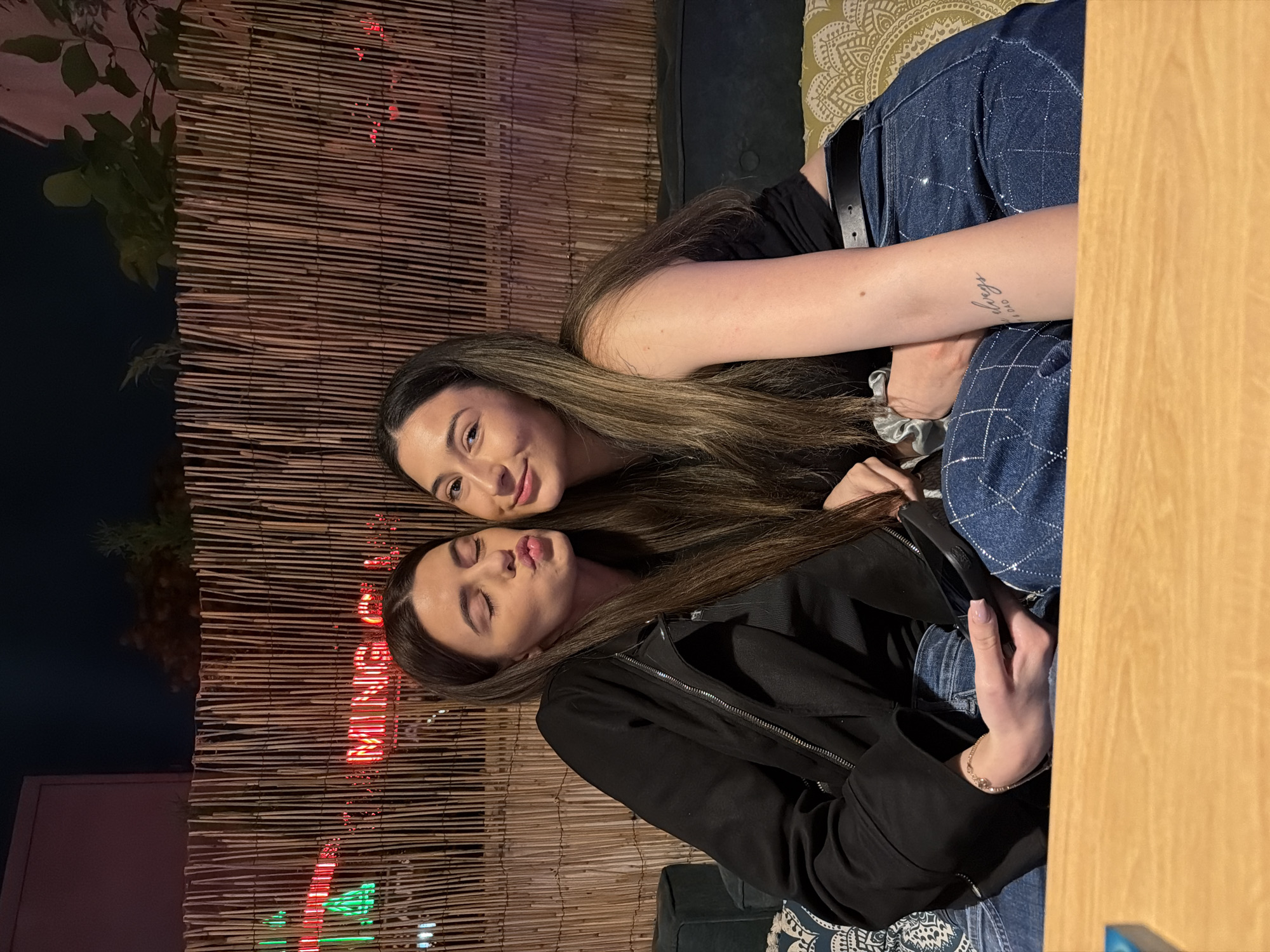 This nighttime image depicts two young adult females seated on a dark green upholstered bench with patterned cushions, positioned behind a light-colored wooden table. The woman on the left, with long dark brown hair, wears a black zippered jacket over a dark top and blue jeans. Her left eye is closed, and her lips are pursed in a kissing gesture. Her right hand, adorned with a light-colored bracelet, rests in her lap, holding a dark, rectangular object. The woman on the right, with long, lighter brown hair, wears a black sleeveless top and blue jeans featuring a subtle checkered pattern, secured by a black belt. She is smiling faintly, looking towards the viewer. A tattoo on her left forearm reads "always" with "M & DAD" below it.
Behind the subjects is a fence constructed from vertical bamboo or reed sticks. Through this fence, a red neon sign is visible, distinctly spelling "MING" and appearing to continue as "MING CHA." A green light source is also present to the left of the neon text. The background is dark, suggesting an evening setting, with dim ambient illumination. Dark foliage and architectural structures are visible against the night sky. Based on the recognizable neon sign, the scene is located at the Ming Cha Bubble Tea Bar in Plovdiv, Bulgaria.