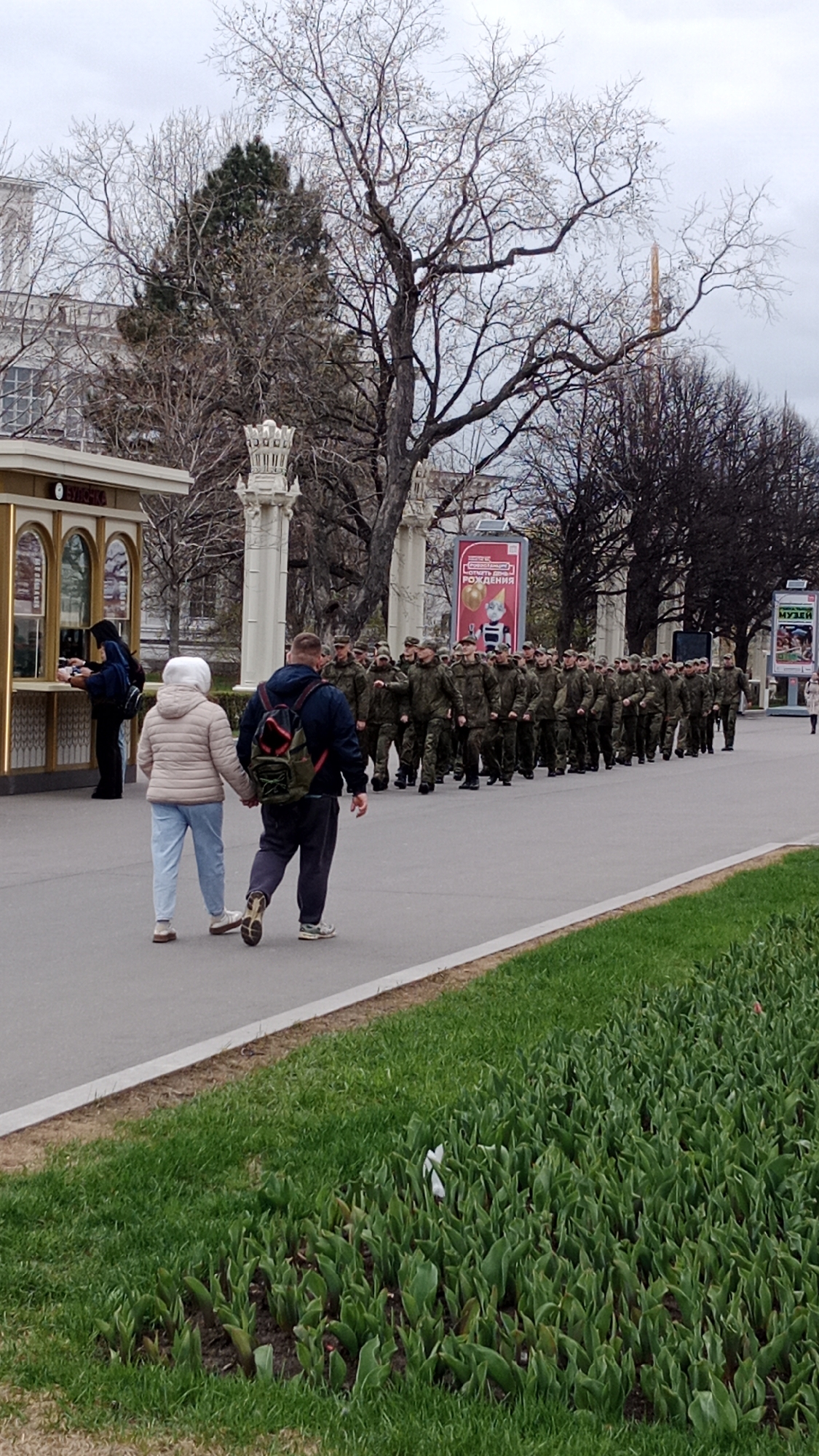 A military parade is underway in Moscow, Russia. A line of soldiers dressed in green camouflage uniforms march in formation along a paved path. Beside the path, an older couple walks hand-in-hand, seemingly observing the parade. In the background, there are buildings, trees with bare branches, ornate pillars, and posters. A kiosk stands to the left, with a person in a dark hoodie being served inside. A grassy area with plants in the foreground adds to the environment. The sky is overcast.