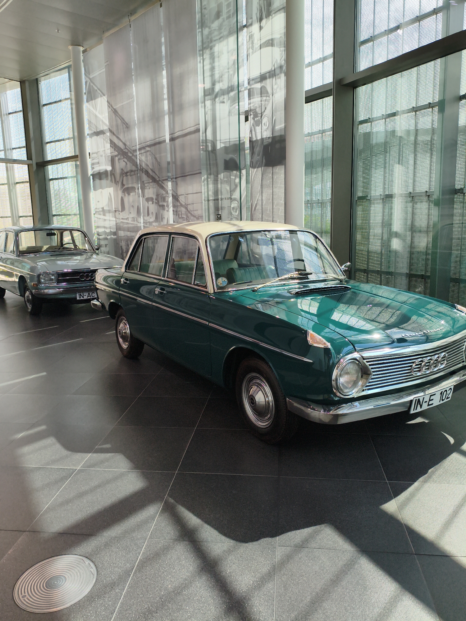 Two classic Audi sedans are displayed in a modern, brightly lit showroom. The car in the foreground is a dark teal sedan with a cream-colored roof, featuring a chrome grille and bumper. Its license plate reads "IN-E 102". Behind it, to the left, is a light blue sedan with a similar design, its license plate reading "IN-AC". The cars are parked on a dark tiled floor that reflects the light from the large windows and the ceiling. The background consists of large glass windows with a grid pattern and sheer white curtains or screens that display subtle, abstract graphics, possibly related to the history of Audi. The overall environment suggests a museum or exhibition space. Given the license plate "IN-E 102" and the presence of classic Audi vehicles, the location is likely the Audi Forum in Ingolstadt, Germany. There are no people visible in the image. The scene is static, showcasing the automobiles in a clean and well-maintained setting.