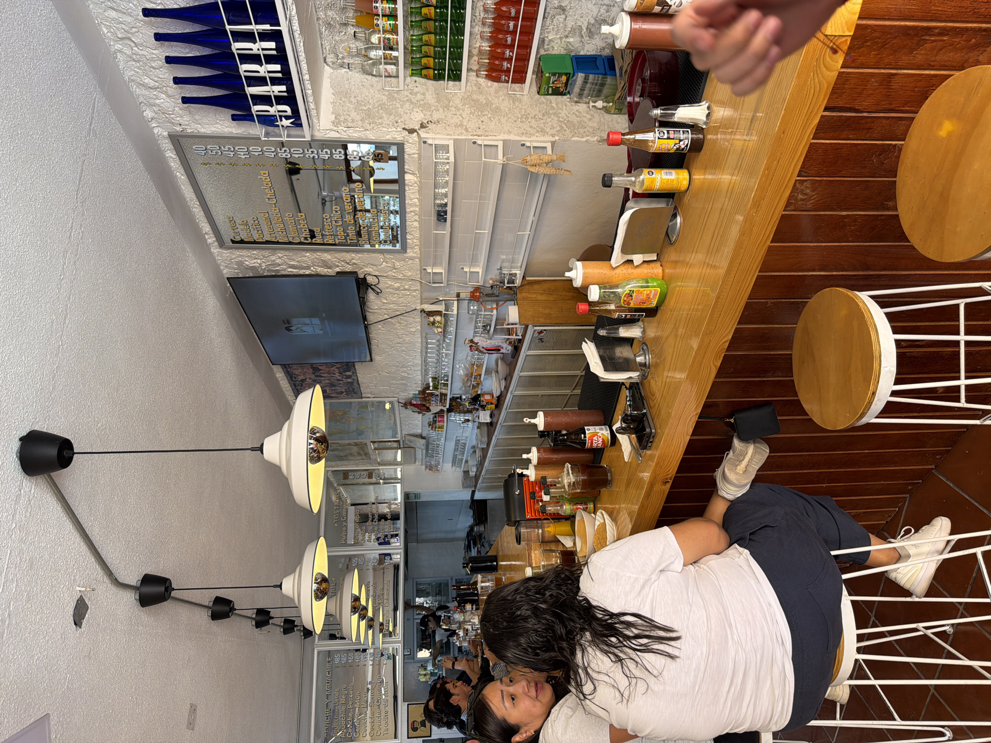 The image depicts the interior of a casual restaurant or bar located in San Andrés Cholula, Mexico. The scene focuses on a wooden bar counter with several patrons seated on white-framed stools. In the foreground, two individuals are positioned at the bar. One person, seen from behind, has long, dark, wavy hair and wears a white t-shirt and dark pants, seated on a stool with their right foot hooked on the rung, a small black pouch hanging from the bar near their left foot. Adjacent to them, another person with dark hair and a white t-shirt faces left, engaged in conversation with a slight smile. Further down the bar to the left, additional patrons are visible.
The bar counter is well-stocked with various condiments, including squeeze bottles of sauces, hot sauce bottles, salt and pepper shakers, and napkin dispensers. Drinks in glasses, including one with a salted rim, are present, alongside small white bowls containing food items. A black mobile phone is visible on the counter. The establishment features white, textured walls. Overhead, a series of black conduit pipes support multiple white, industrial-style pendant lights with reflective interiors.
Along the left wall, two framed menus in Spanish list "Ceviche y Aguachile" options and prices. On the right wall, a larger framed menu in Spanish displays "Cerveza," "Refresco," and "Michelada" prices. Above this menu, a flat-screen television shows an image. Below the TV and menus, white shelving units are mounted on the wall. To the right, shelves display numerous blue bottles arranged to spell "BARRI," along with rows of bottled beverages in green, red, and clear glass. A small wooden decorative item resembling dried fish hangs on one of the central white shelves. The overall ambiance is that of a bustling, informal eatery.