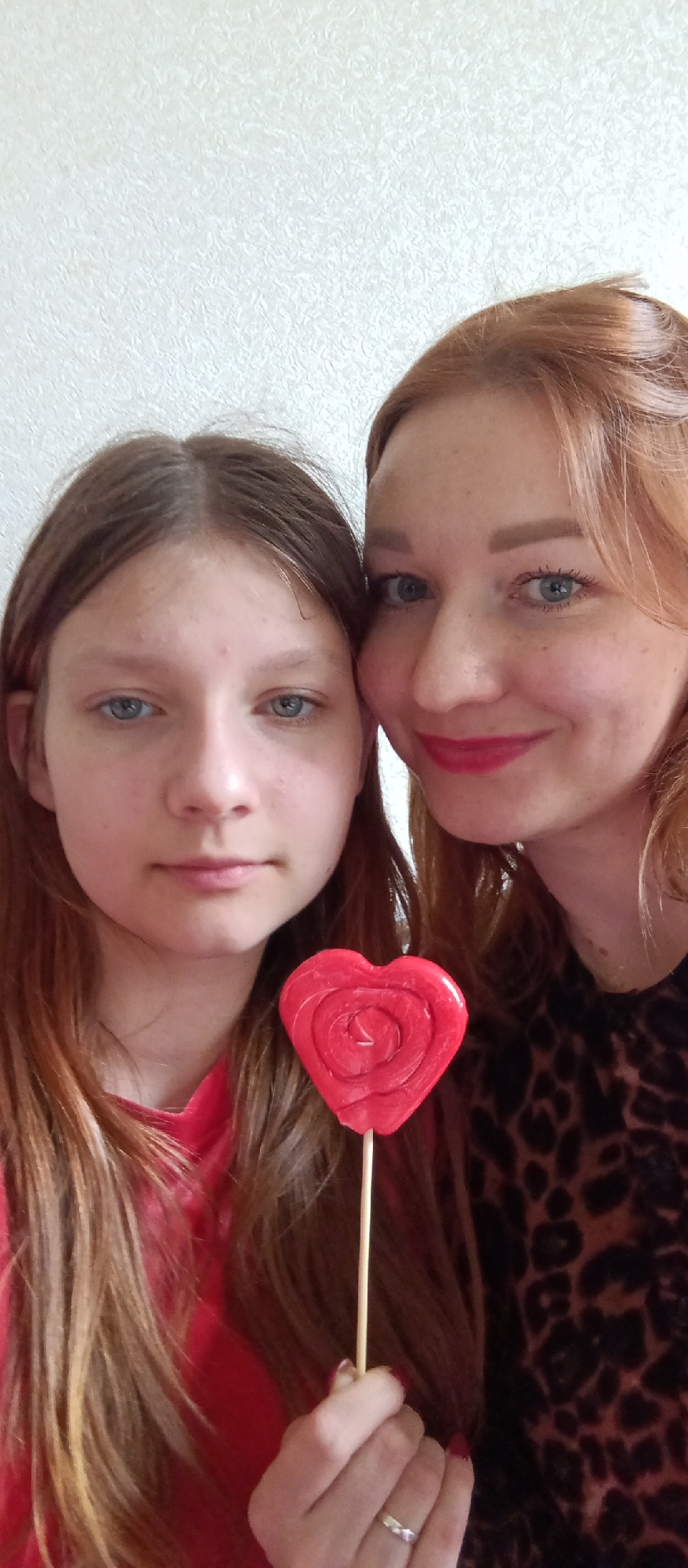 A close-up, vertical portrait captures two females posing together indoors. On the left is a younger female with long, straight brown hair parted in the middle, blue eyes, and fair skin, wearing a red shirt. Her expression is neutral as she looks directly at the camera. To her right, partially visible, is an older female with auburn hair styled up, blue eyes, fair skin with noticeable freckles, and red lipstick, smiling warmly at the camera. She is wearing a dark top with a leopard print pattern.
In the foreground, held by the younger female's left hand, is a vibrant red, heart-shaped lollipop with a spiral design, positioned centrally between the two subjects. A silver ring with a small embellishment is visible on the younger female's ring finger. The background is a plain, light-colored (likely white or off-white) textured wall, suggesting an indoor setting. The lighting is soft and even, without harsh shadows. There is no visible text. The interaction of the subjects and the heart-shaped lollipop suggests a moment of affection, possibly for Valentine's Day or a similar occasion.