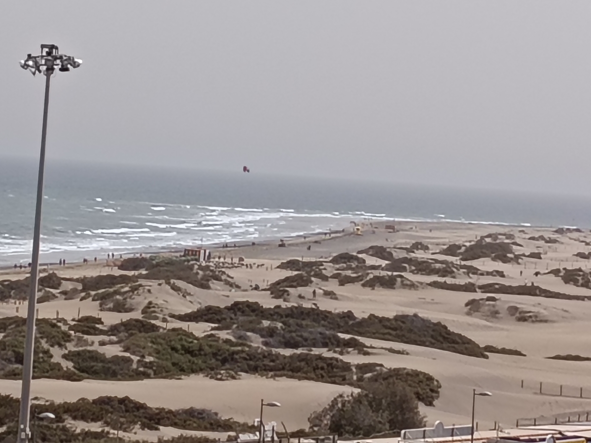 The image depicts a coastal scene in San Bartolomé de Tirajana, Spain. In the foreground, sandy dunes are covered with vegetation. A tall light pole stands prominently on the left side of the frame. In the middle distance, a beach is visible with people walking along the shoreline, and waves are crashing on the shore. A small red object is visible in the sky, possibly a kite or a balloon. Several small buildings or structures are visible along the beach. In the background, the ocean stretches to the horizon under a cloudy sky. Streetlights and railings can be seen in the lower right corner, indicating an urban area near the beach.