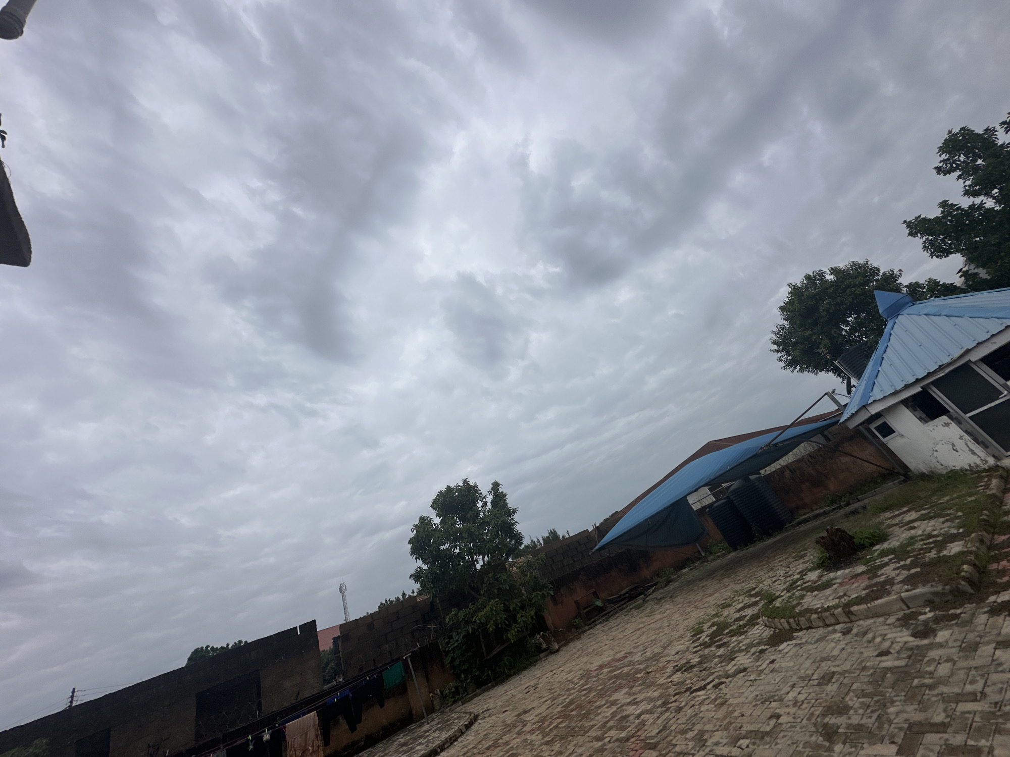 The image captures an overcast sky above a residential area in Jos, Nigeria. In the foreground, a patterned brick pathway extends towards a low, dark-colored brick wall with some barbed wire along its top edge. Clothes are hung out to dry on a line attached to the wall, with several dark-colored garments visible. To the right of the pathway, a building with a blue, corrugated metal roof and white walls is partially visible, with some windows and a door. Further in the background, more brick structures and trees are present. A tall communication tower can be seen in the distance to the left. The overall scene suggests a somewhat weathered urban environment under a gloomy sky.