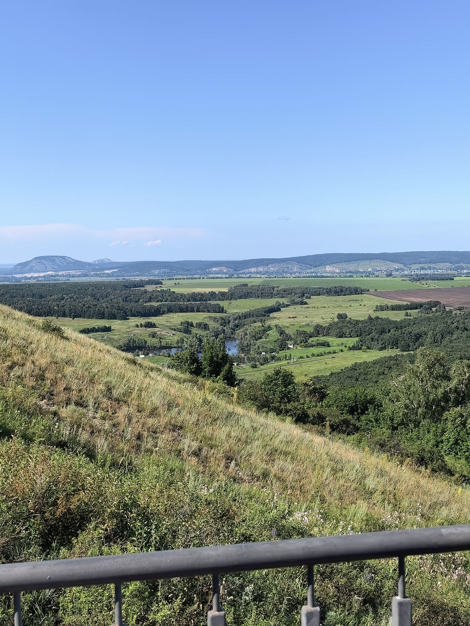 The image shows a panoramic view of a rural landscape under a clear blue sky. In the foreground, a grassy slope descends, with a metal railing along the bottom edge. The midground features a vast expanse of green fields, interspersed with patches of trees and a winding river or lake. In the distance, a range of rolling hills forms the horizon. The overall scene depicts a serene and natural environment, possibly in the vicinity of Akhmerovo, Russia, given the provided context. There are no people or explicit actions depicted in the image.
