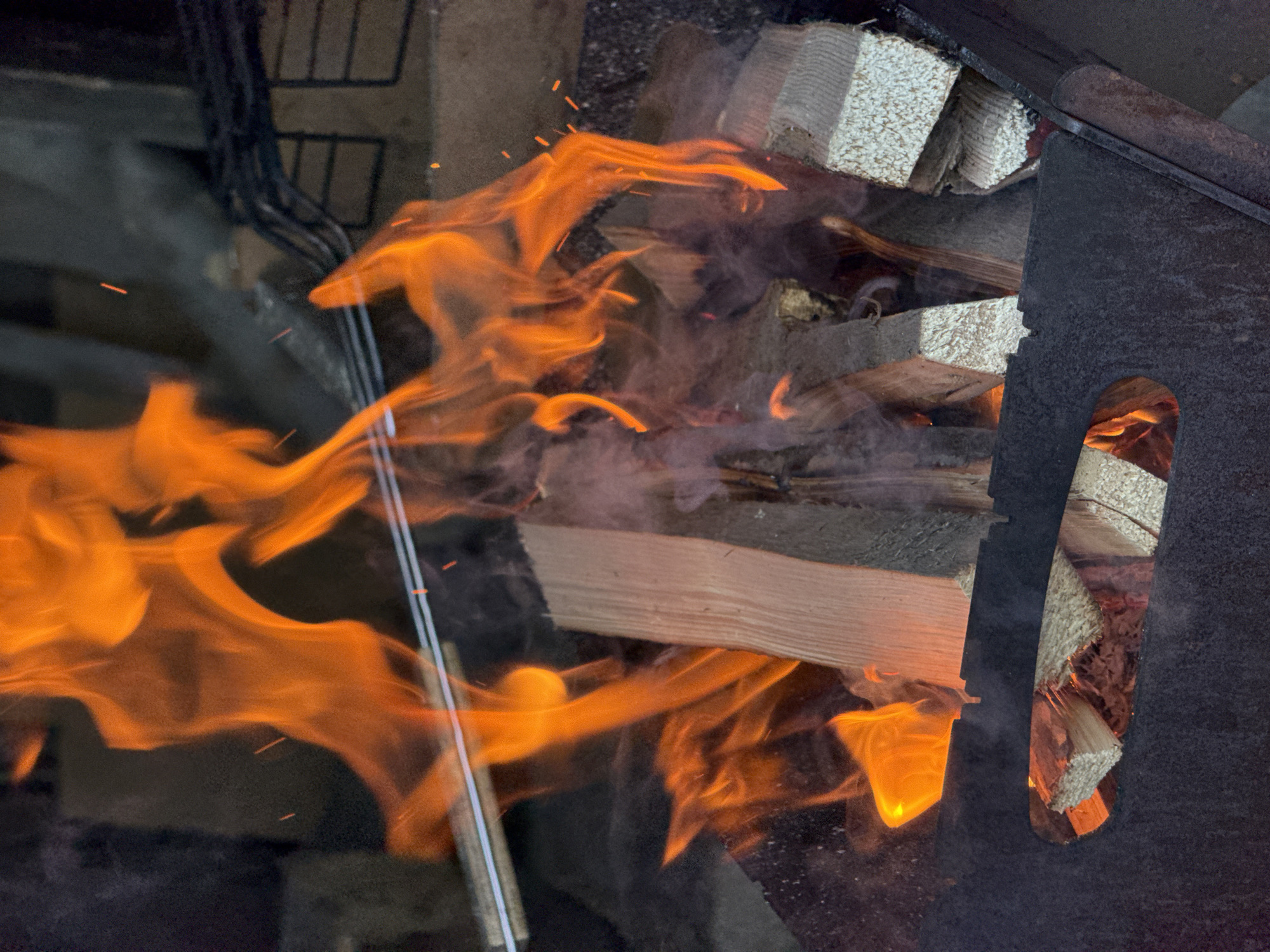 The image shows a wood-burning stove in operation. Flames are visible within the stove's firebox, and smoke is present. The stove appears to be loaded with wood. A metal grate or other fixture is partially visible behind the flames. The surrounding environment is dark and indistinct. There is no indication in the image itself that the location is in Зыково, Russia.