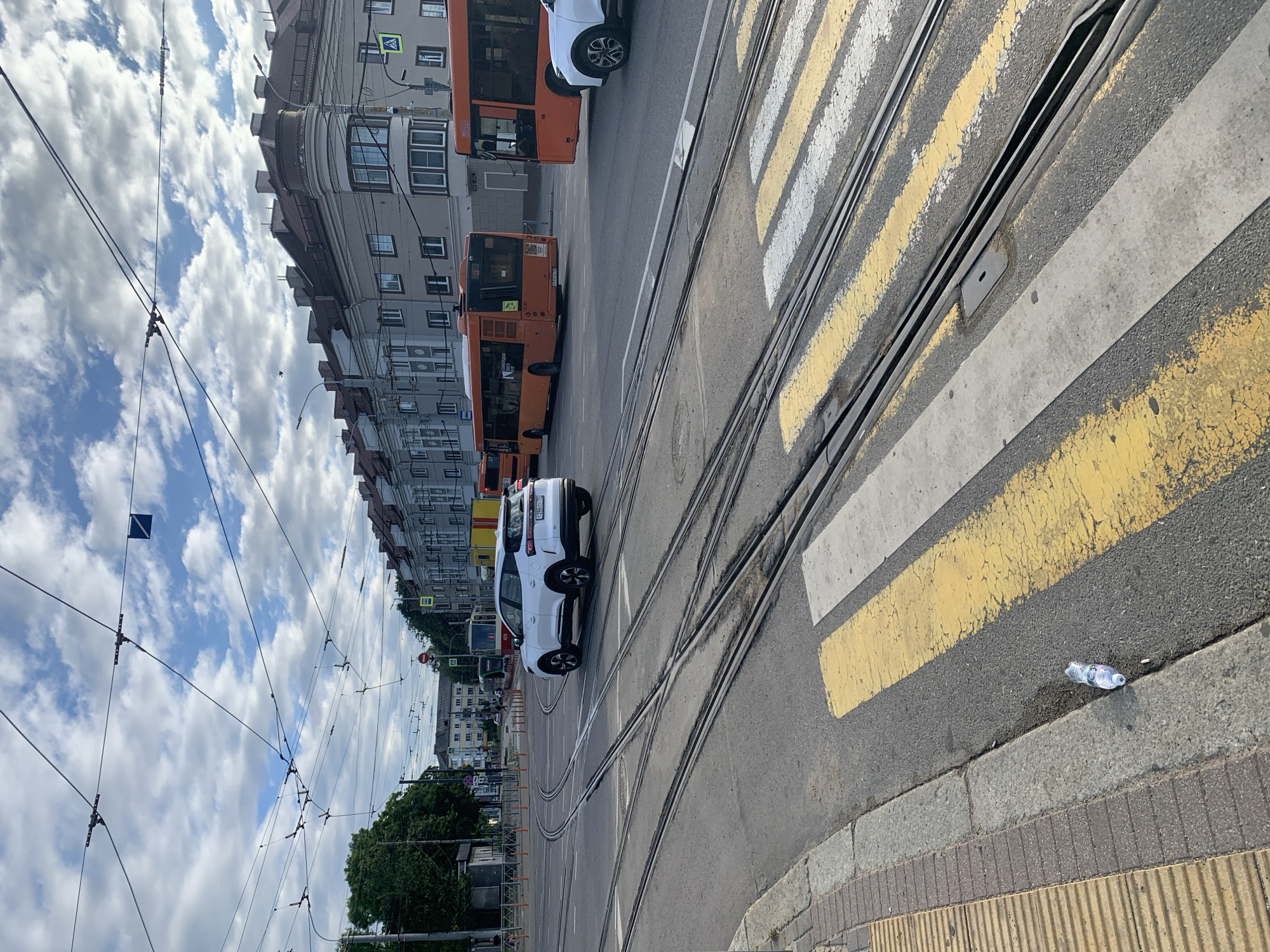 The image captures a street scene in an urban environment, possibly in the city of Unknown, Unknown. In the foreground, a white SUV is turning at an intersection, with tram tracks clearly visible on the asphalt. A pedestrian crossing with yellow and white stripes is also prominent. Several orange buses are driving on the road, with one appearing to be stopped or moving slowly. In the background, buildings with multiple stories and peaked roofs line the street, with a cloudy sky overhead. Overhead wires, likely for the trams, crisscross the sky. A discarded plastic water bottle is on the ground near the curb.
