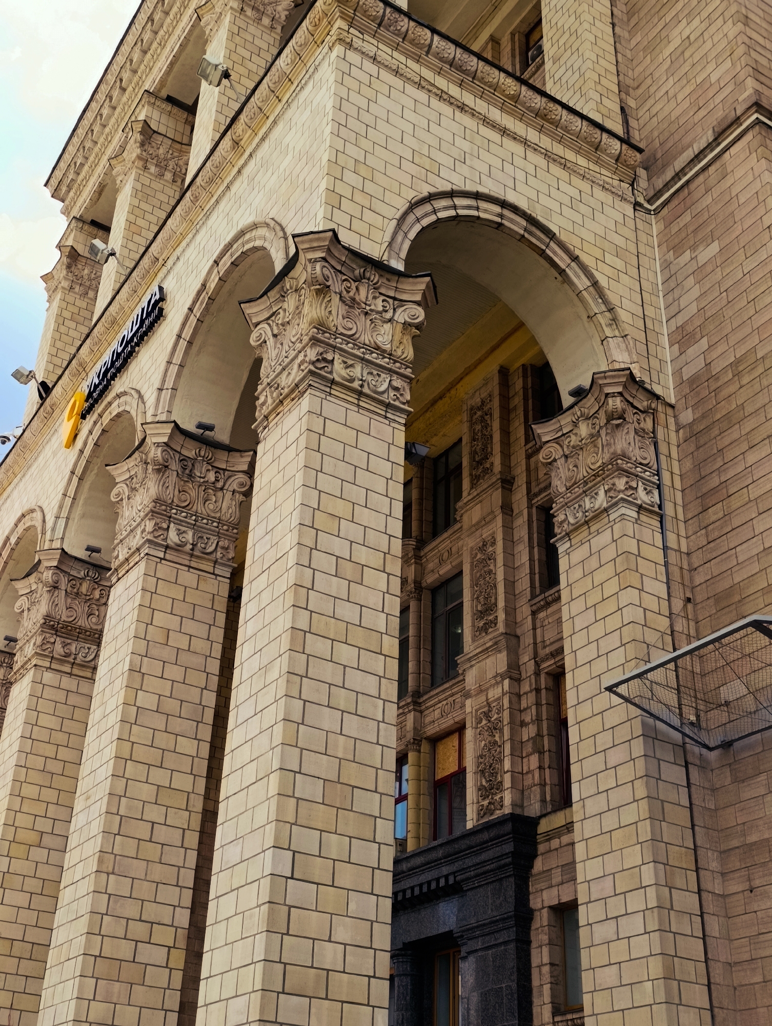 The image shows the facade of a grand building in Kyiv, Ukraine, specifically the Central Post Office building, also known as Ukrposhta. The perspective is from below, looking upwards at the building's ornate architecture. The building is constructed of light-colored bricks and features large arches supported by intricately carved columns. Above the arches, there are decorative moldings and windows. A sign with yellow lettering and a yellow "9" symbol is visible above one of the arches. The sky is visible in the upper left corner, appearing overcast. The detail of the stonework and brickwork suggests a building of historical significance and impressive craftsmanship.