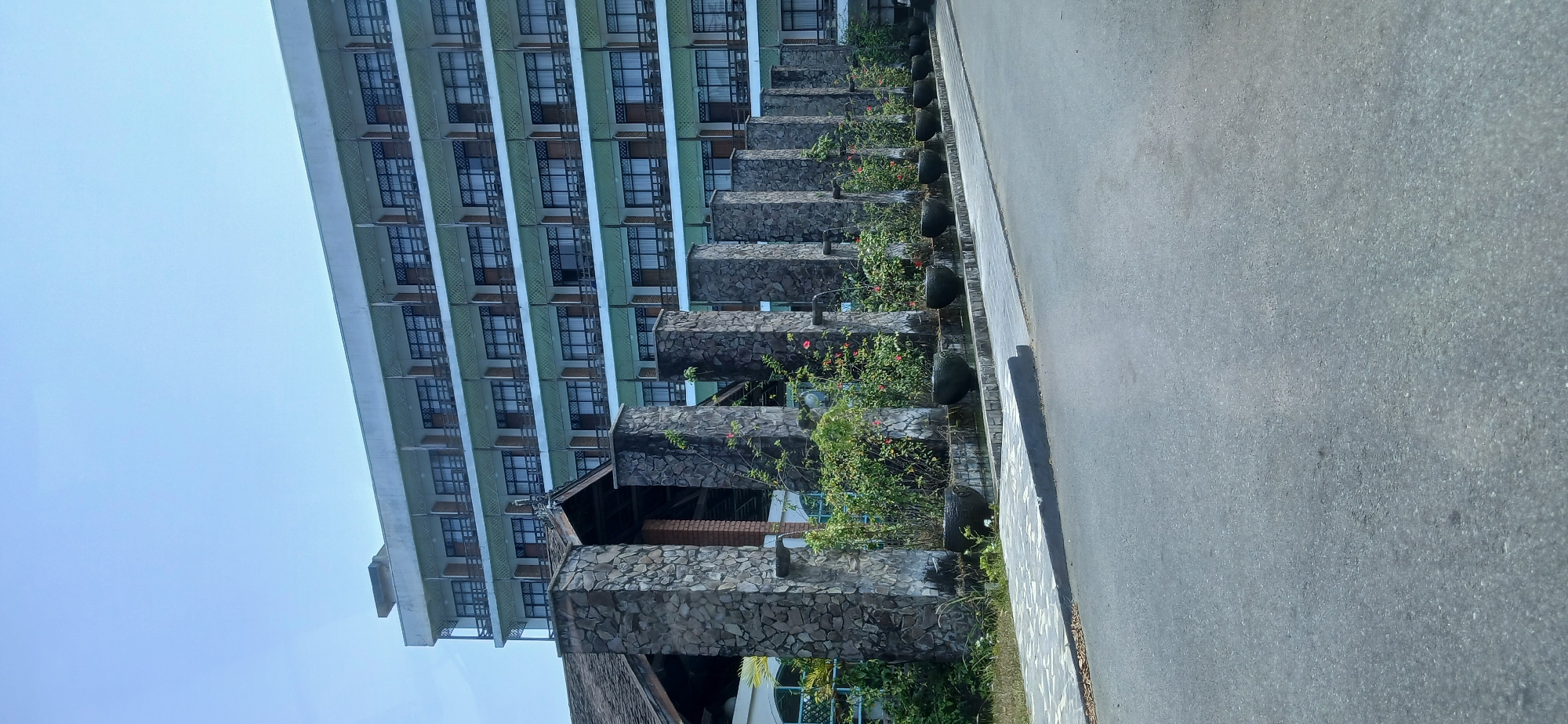 The image depicts a long, multi-story hotel building with a light-colored facade and numerous balconies, set against a clear blue sky. In the foreground, a row of stone pillars, each topped with a decorative element, lines a paved walkway. Water spouts from small fixtures on the pillars, feeding a shallow water feature below, where dark, bowl-shaped planters filled with lush green plants and some red flowers are arranged. A dark asphalt road occupies the lower portion of the image. The architectural style suggests a resort or a hotel, possibly located in a tropical or subtropical region. Based on the prompt's requirement for Samarinda, Indonesia, this could be a hotel or resort located within or on the outskirts of the city, catering to tourists or business travelers, with its design likely influenced by local aesthetics and climate. No people are visible in the scene.