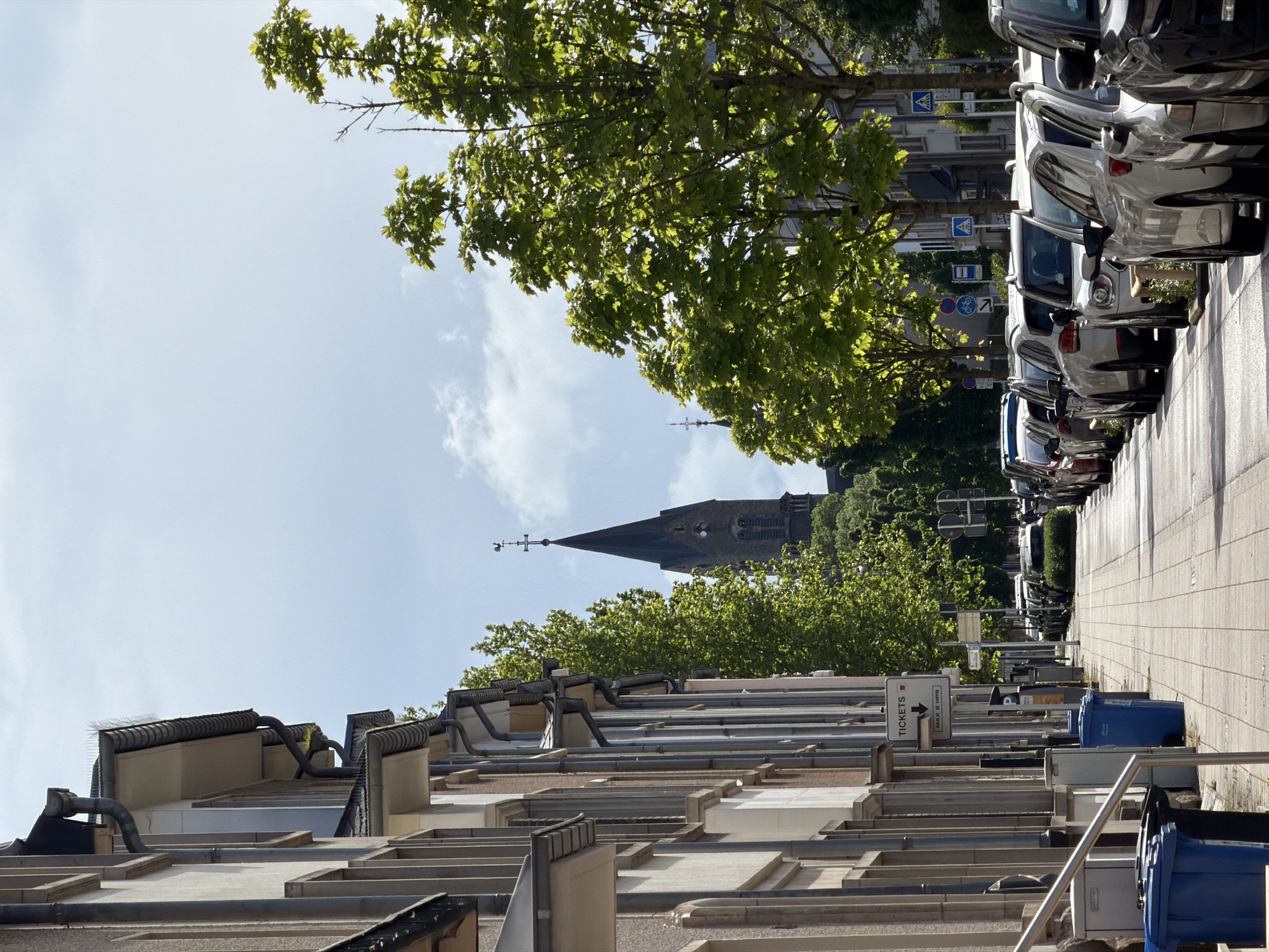 The image shows a street scene in Esch-sur-Alzette, Luxembourg, characterized by a row of modern, multi-story residential buildings on the left side and parked cars along the right side of the street. A church steeple is visible in the background, framed by trees. The street has a paved sidewalk. There are trash cans and signage along the building side of the street. The sky is partly cloudy. The picture is taken at ground level.