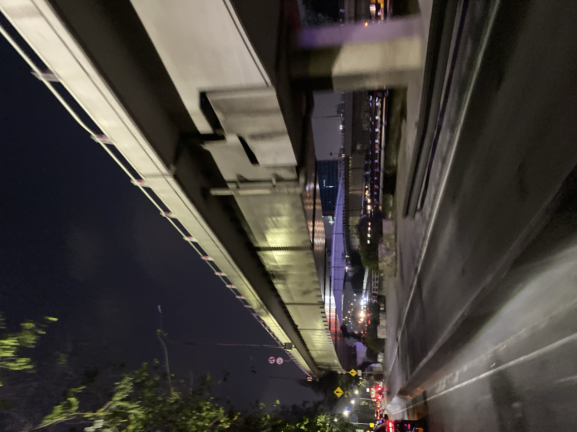 The image depicts a nighttime view of an elevated highway structure in Tangerang, Indonesia. The highway, made of concrete and metal, is illuminated by streetlights, casting a warm glow on its underside. Below the elevated highway, another road or overpass can be seen, also with lights from vehicles. On the left side of the image, trees with green leaves overhang the scene. The road in the foreground shows signs of being wet, possibly from recent rain, with a lane marked by white lines. Several cars are visible on the road, their headlights and taillights creating streaks of light. Yellow warning signs are posted along the roadside, indicating potential hazards. The sky is dark and cloudy, typical of a city at night. The overall impression is one of urban infrastructure and nighttime traffic.